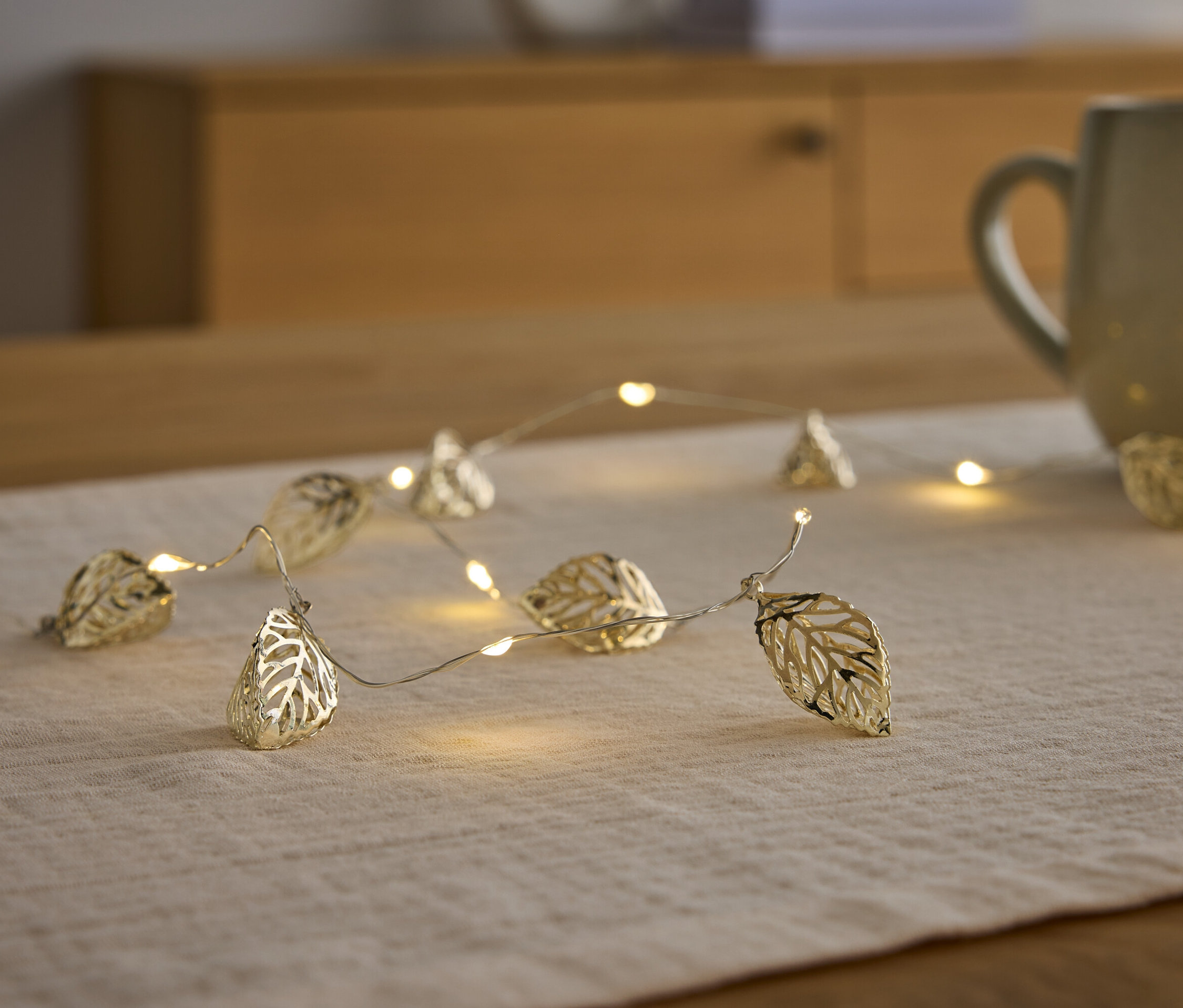 Arany színű, levél alakú fényfüzér fekszik egy asztali futón egy bögre mellett.