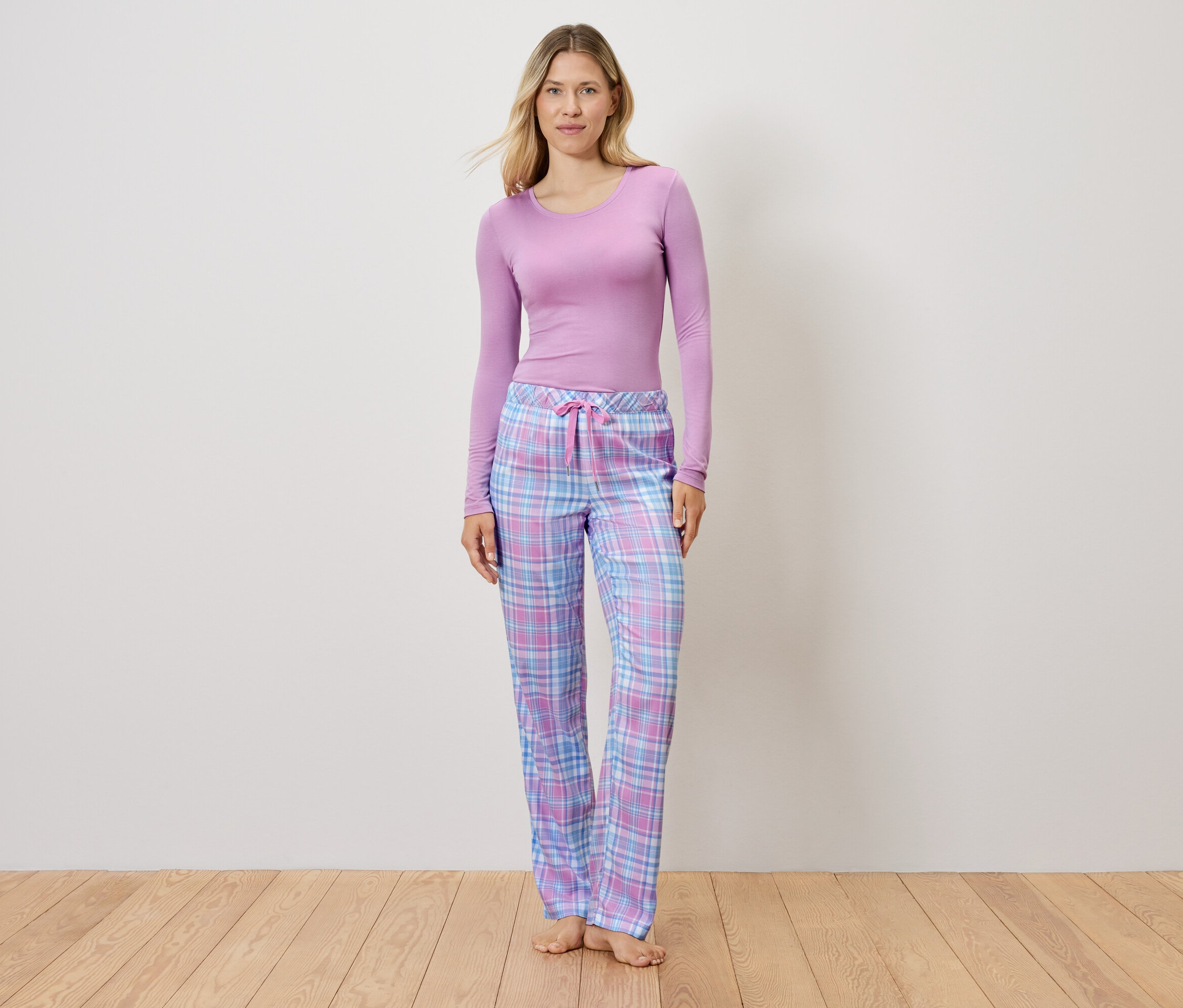 Lila felsőt és kockás pizsamanadrágot viselő nő áll a fapadlón.