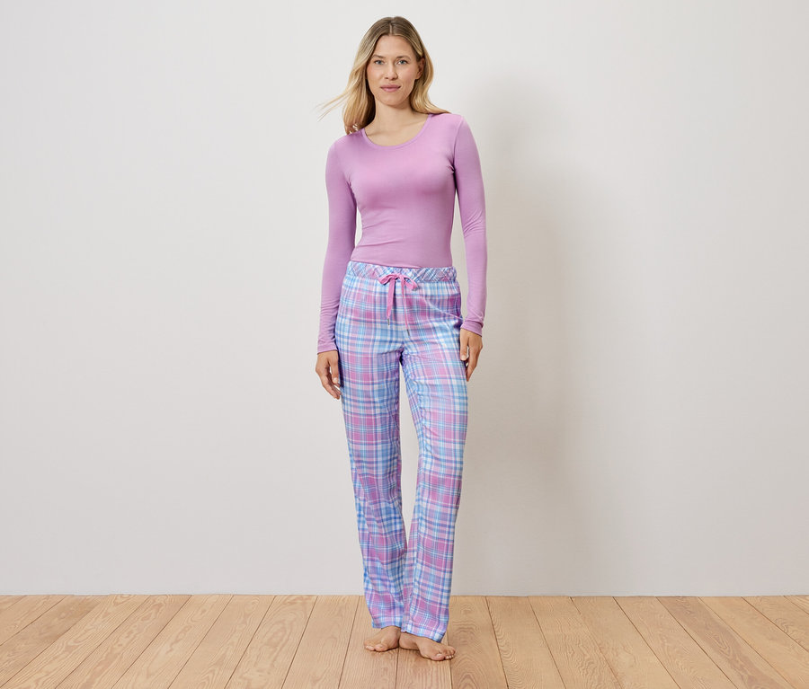 Lila felsőt és kockás pizsamanadrágot viselő nő áll a fapadlón.