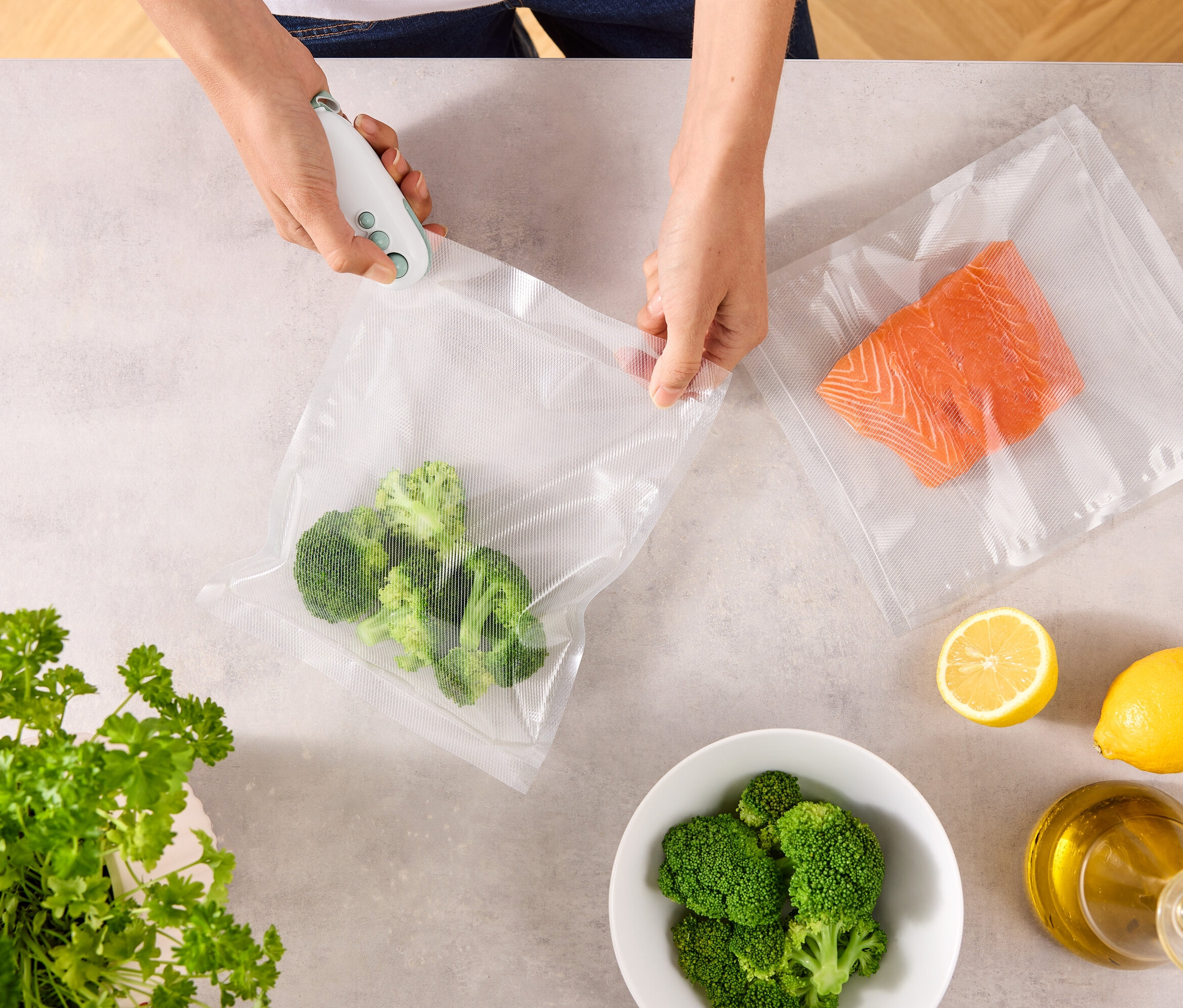 Egy személy vákuumozóval zár le egy zacskót brokkolival. Az asztalon van még egy zacskó lazaccal, citromok, egy tál brokkoli, petrezselyem és egy üveg olaj.