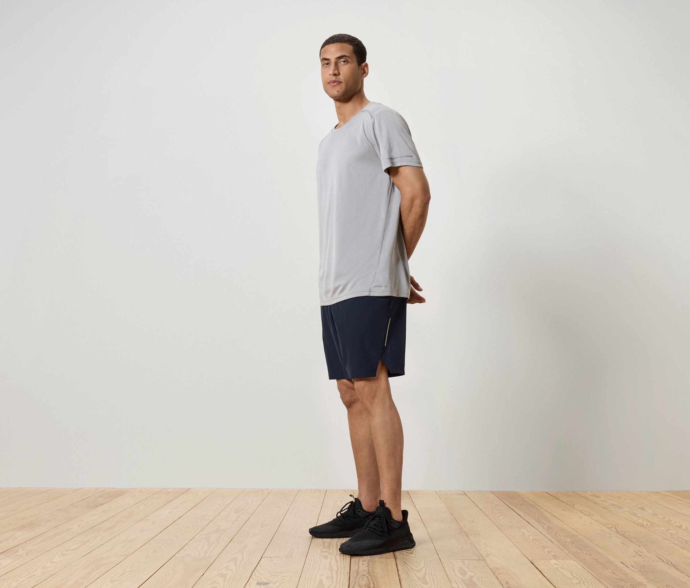 Egy férfi oldalra állva, szürke pólót, sötétkék rövidnadrágot és fekete tornacipőt visel.
