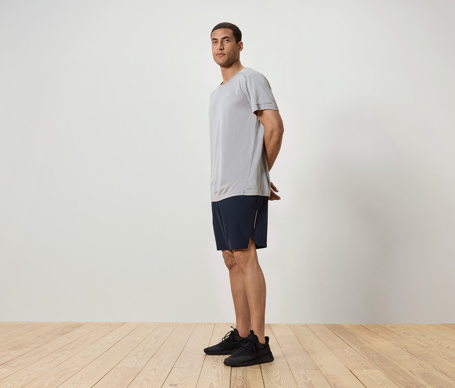 Egy férfi oldalra állva, szürke pólót, sötétkék rövidnadrágot és fekete tornacipőt visel.