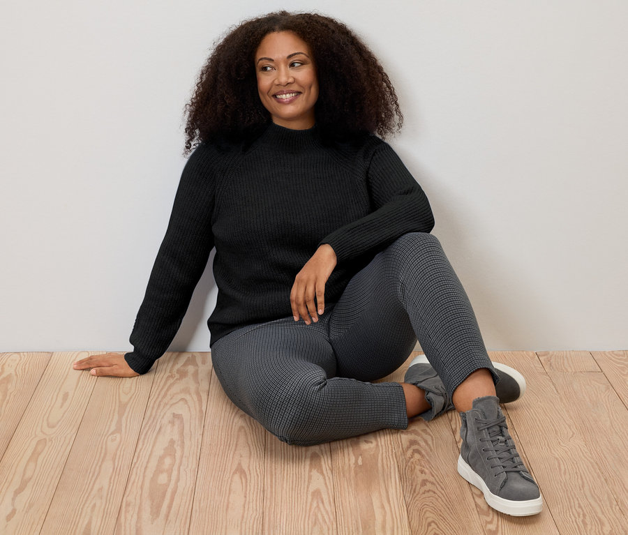 Nő ül a padlón, fekete, laza kötésű, állógalléros pulóvert visel.