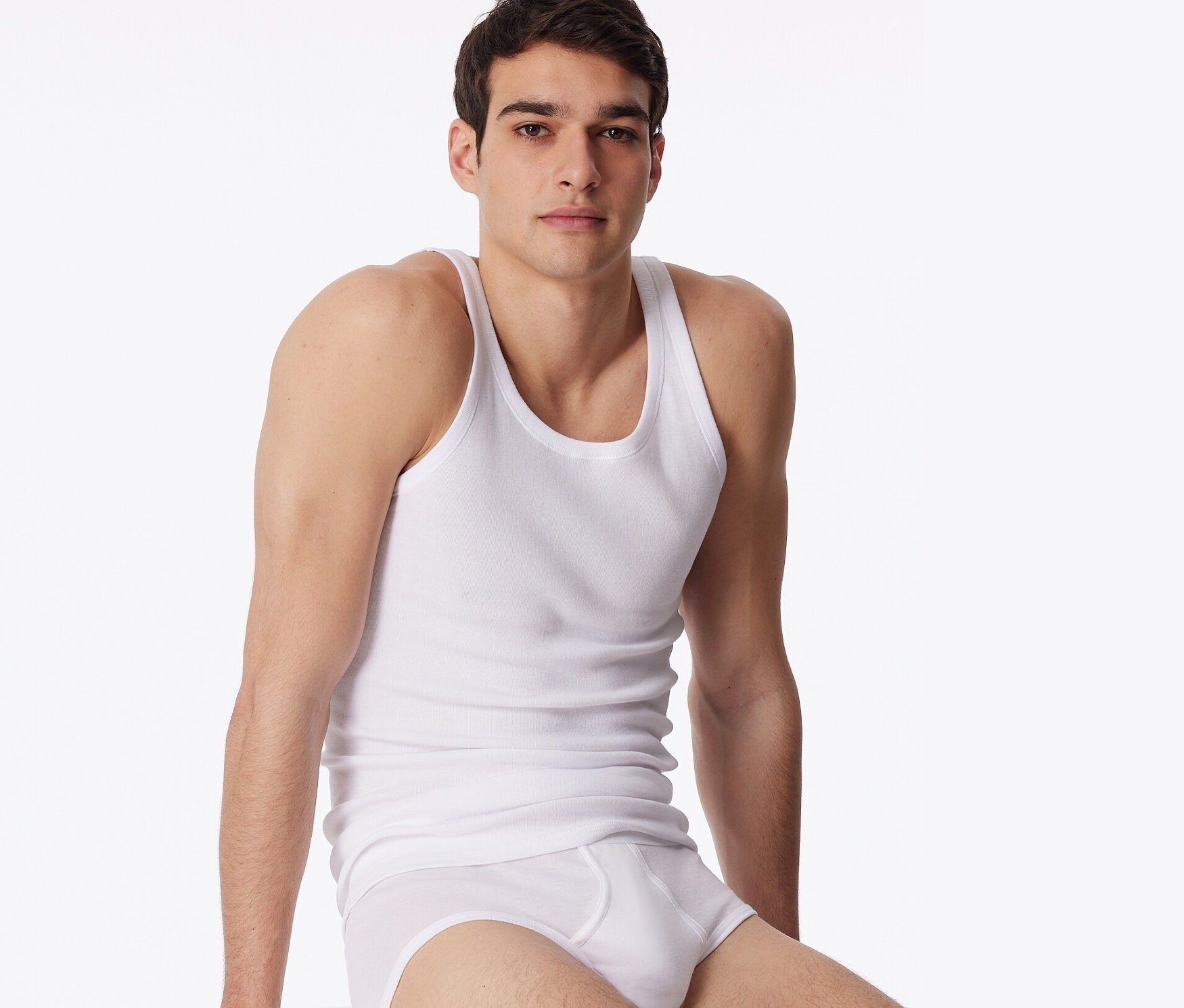 Férfi ül egy fehér kockán, fehér alsónadrágot és 2 db fehér, finoman bordázott SCHIESSER trikót visel.