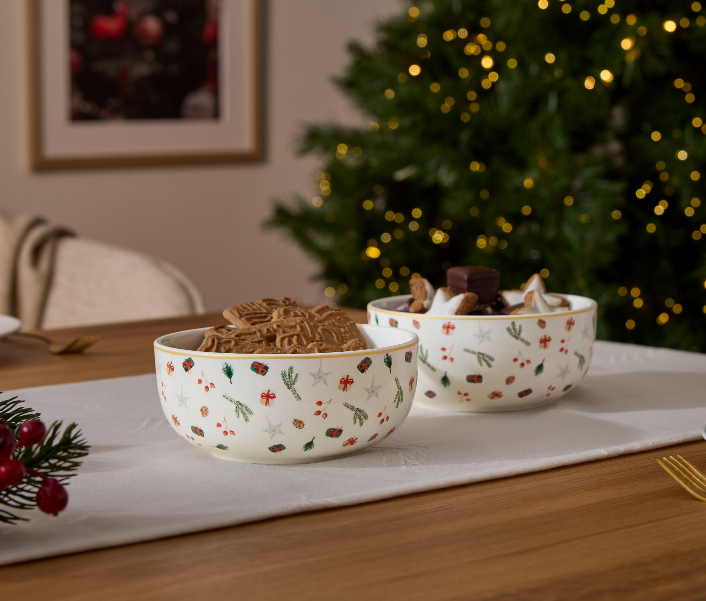 Két karácsonyi mintás tál, az egyik mézeskaláccal, a másik csokoládéval és csillagokkal töltve, egy karácsonyi dekorációval ellátott asztalon.