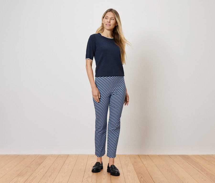 Egy nő áll a fapadlón, sötétkék pólót és kék-fehér csíkos nadrágot visel.