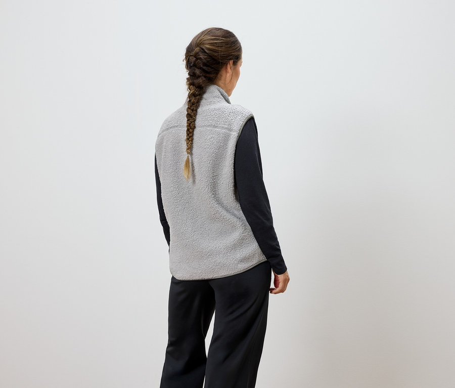 Hátulról látható nő fonott hajjal, szürke mellényt és fekete nadrágot visel.