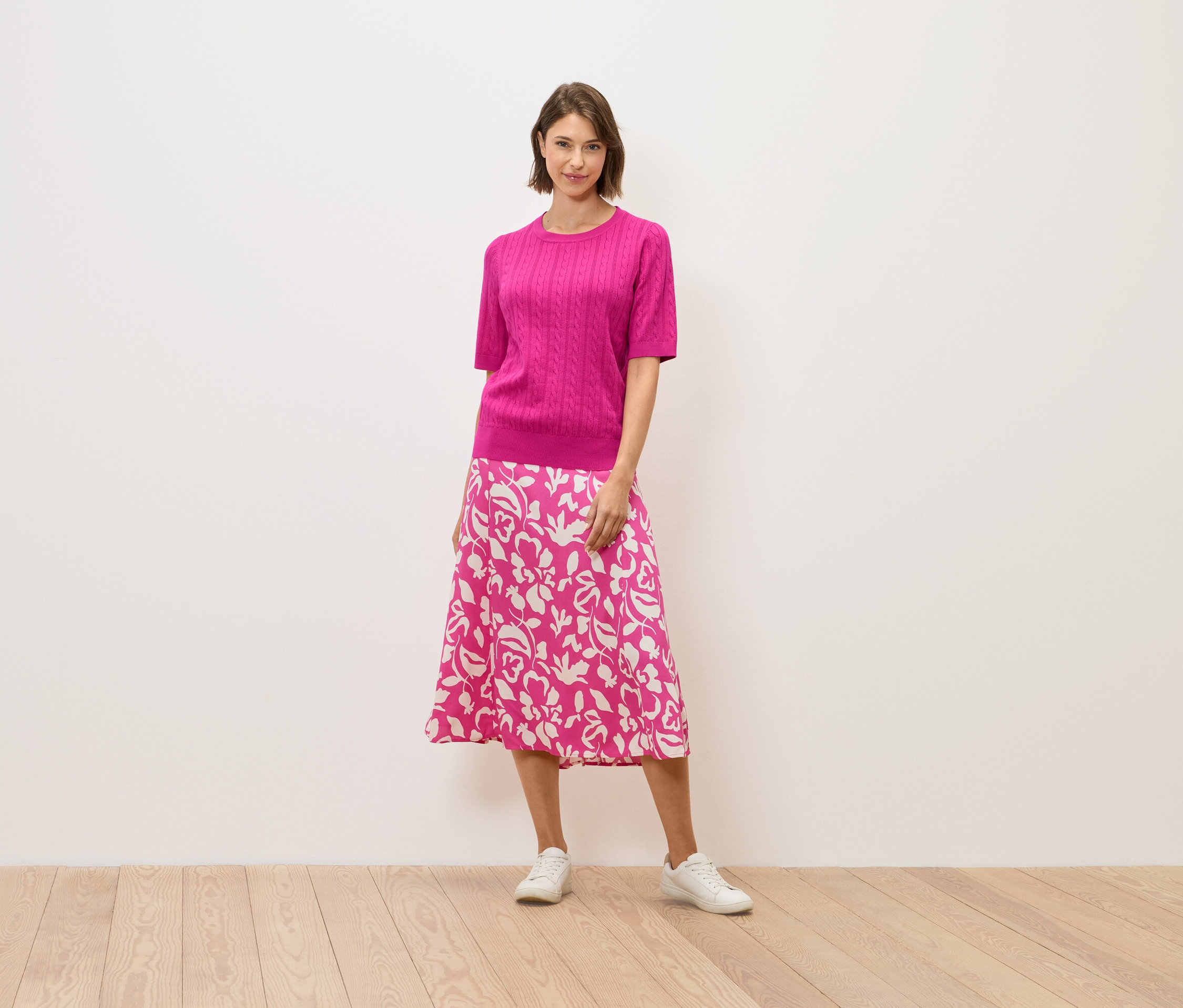 Egy nő rózsaszín pulóverben és virágmintás szoknyában áll egy fapadlón.