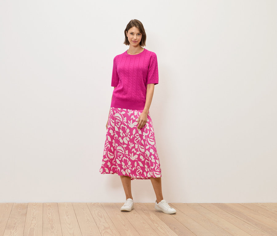 Egy nő rózsaszín pulóverben és virágmintás szoknyában áll egy fapadlón.