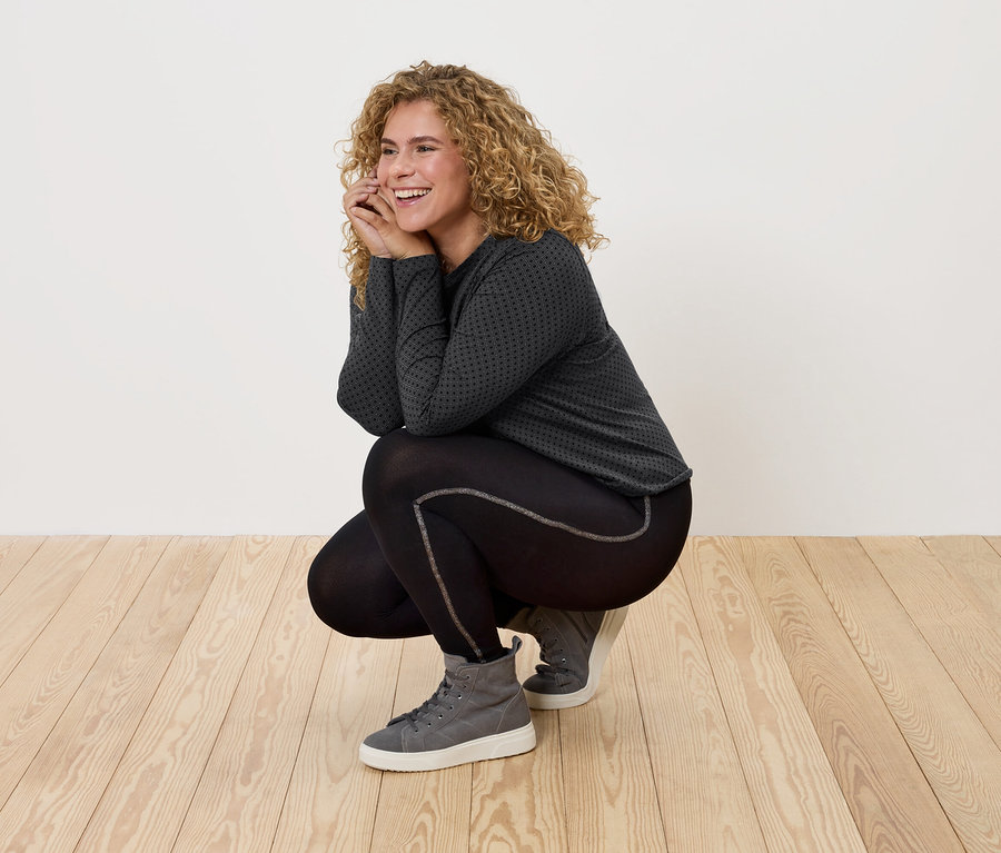 Gyakorlóruhát és szürke tornacipőt viselő, göndör hajú nő pózol a fa padlózaton.