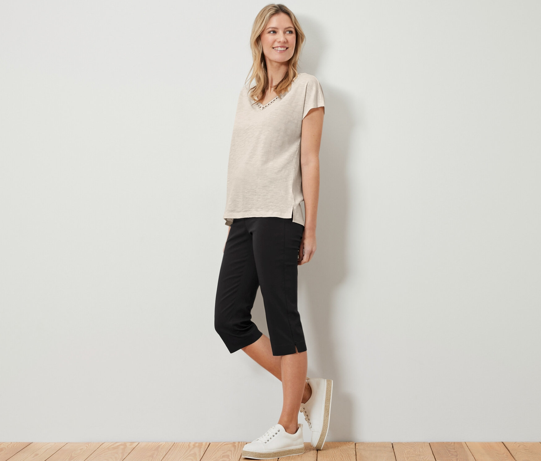Egy nő áll, bézs pólót és fekete 3/4-es sztreccsnadrágot visel.
