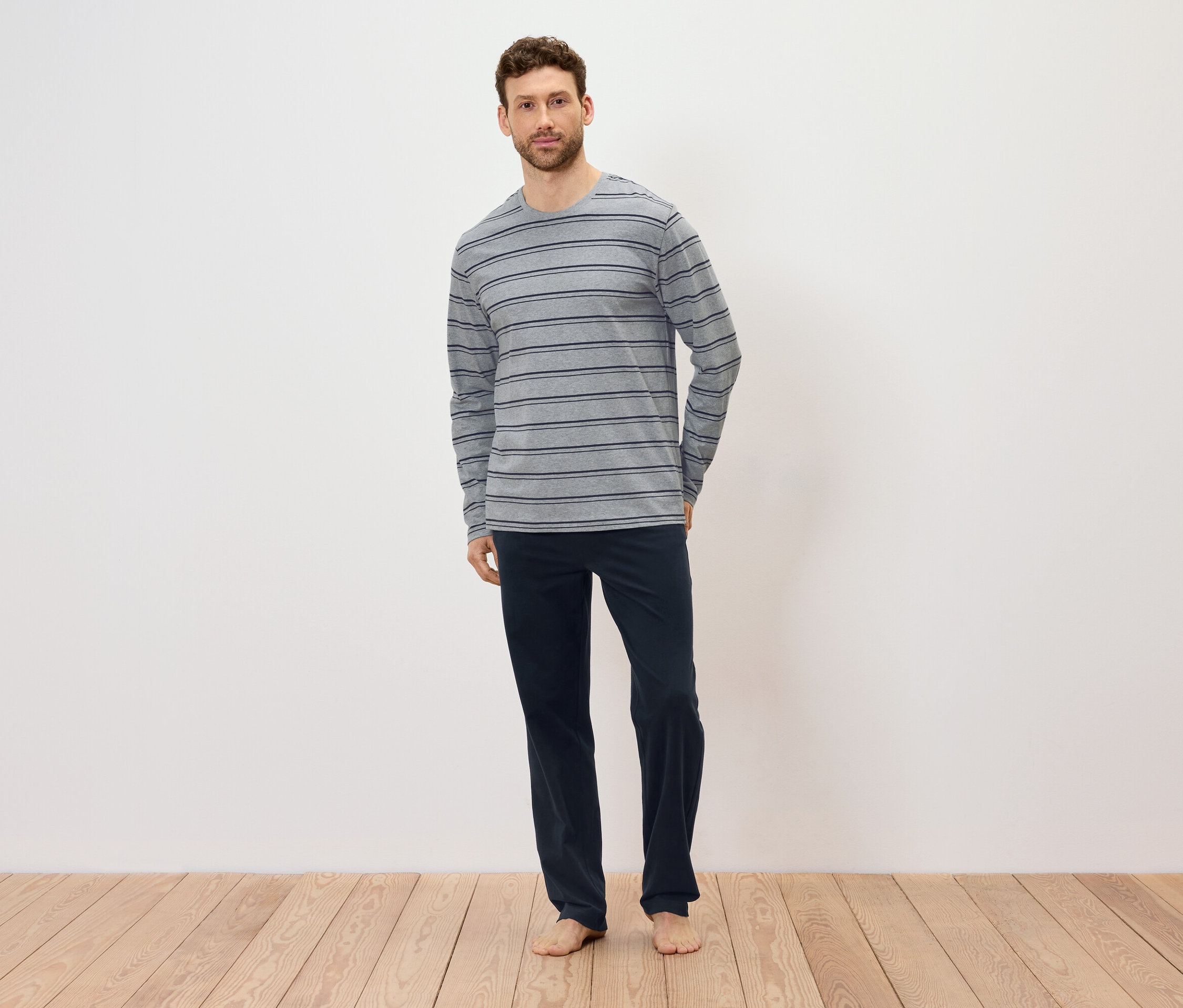 Egy férfi áll a fapadlón, szürke, csíkos, hosszú ujjú inget és sötétkék nadrágot visel.