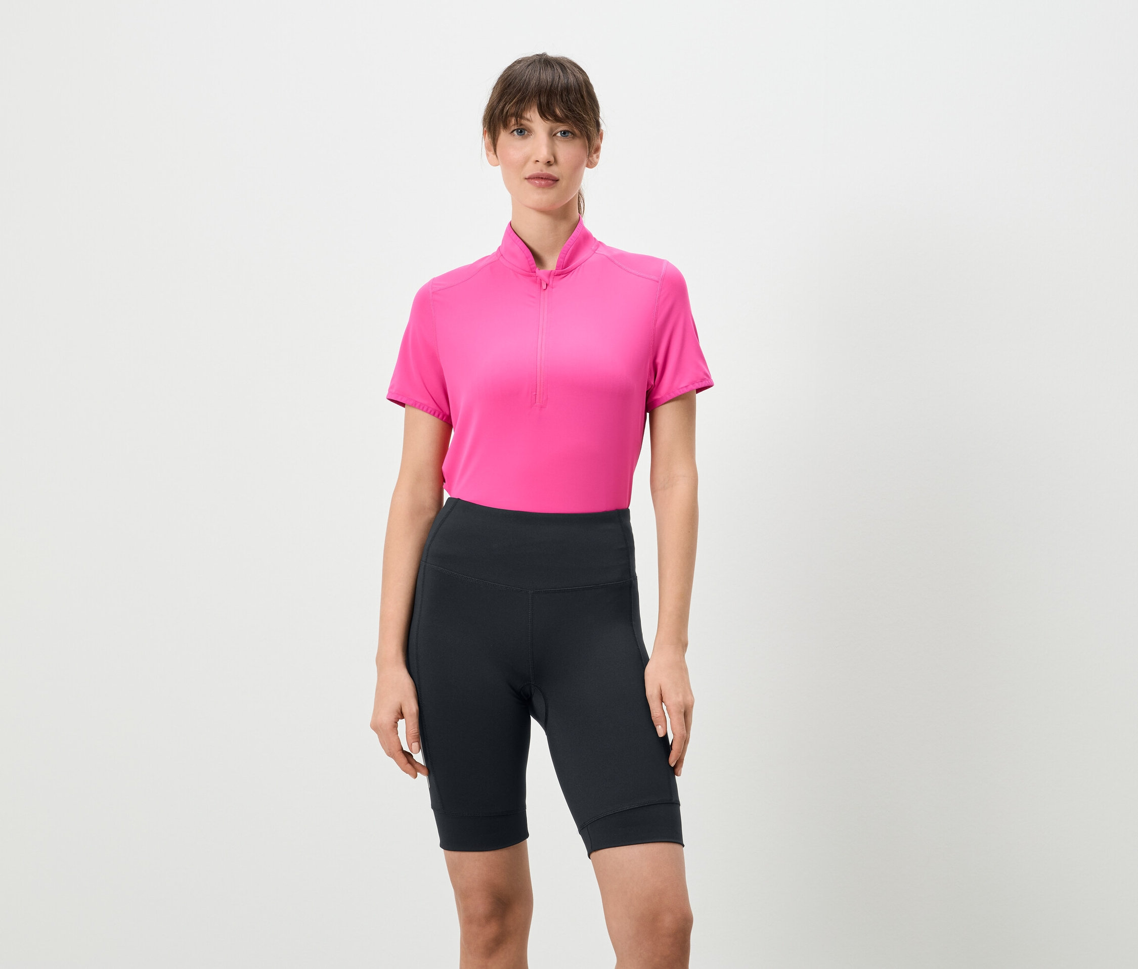 Nő áll fehér háttér előtt, rövid cipzáras rózsaszín pólót és rövid, sötét leggingset visel.
