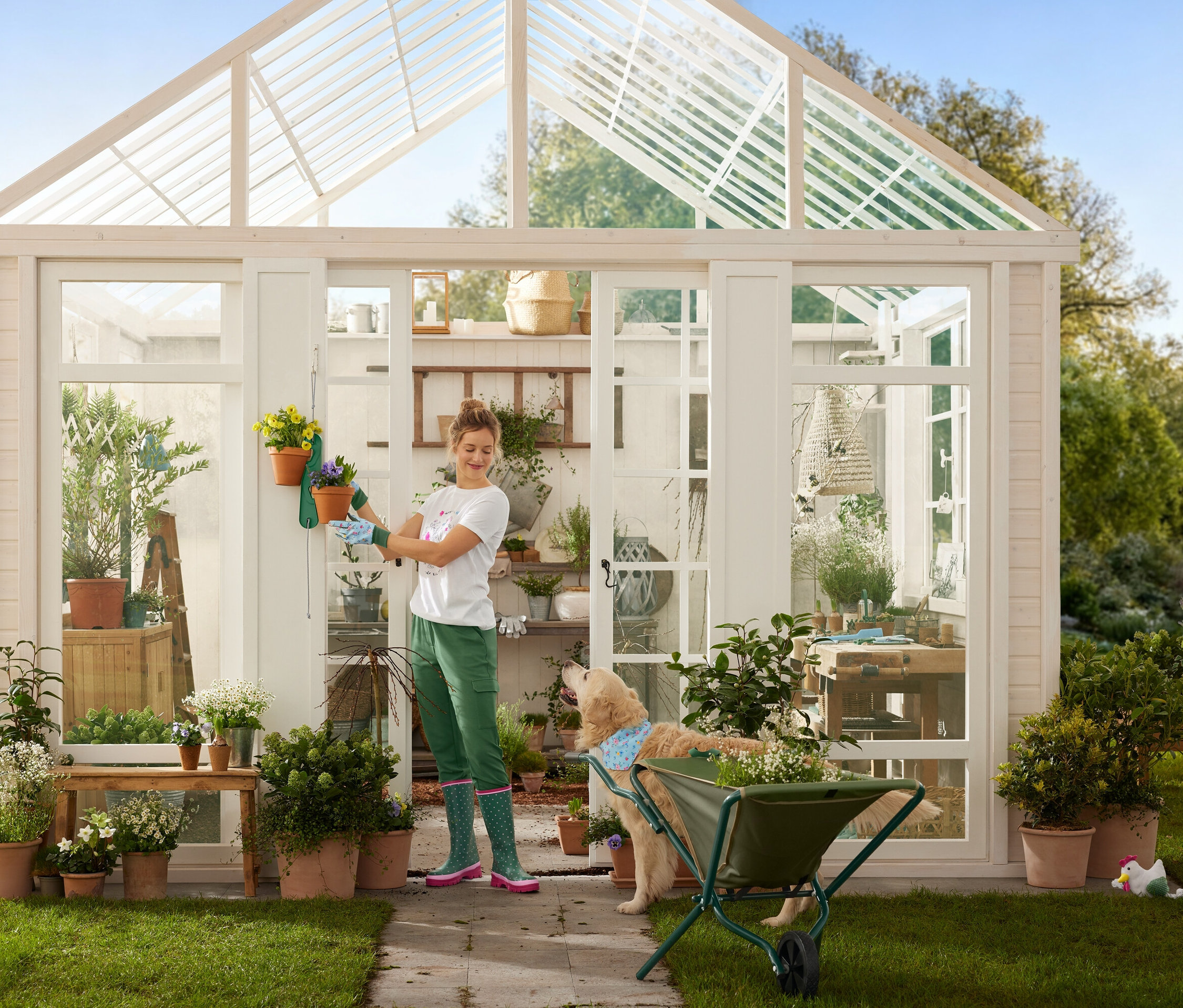 Egy nő kertészkesztyűben virágcserepeket akaszt egy üvegházra, miközben egy kutya egy talicska mellett áll.