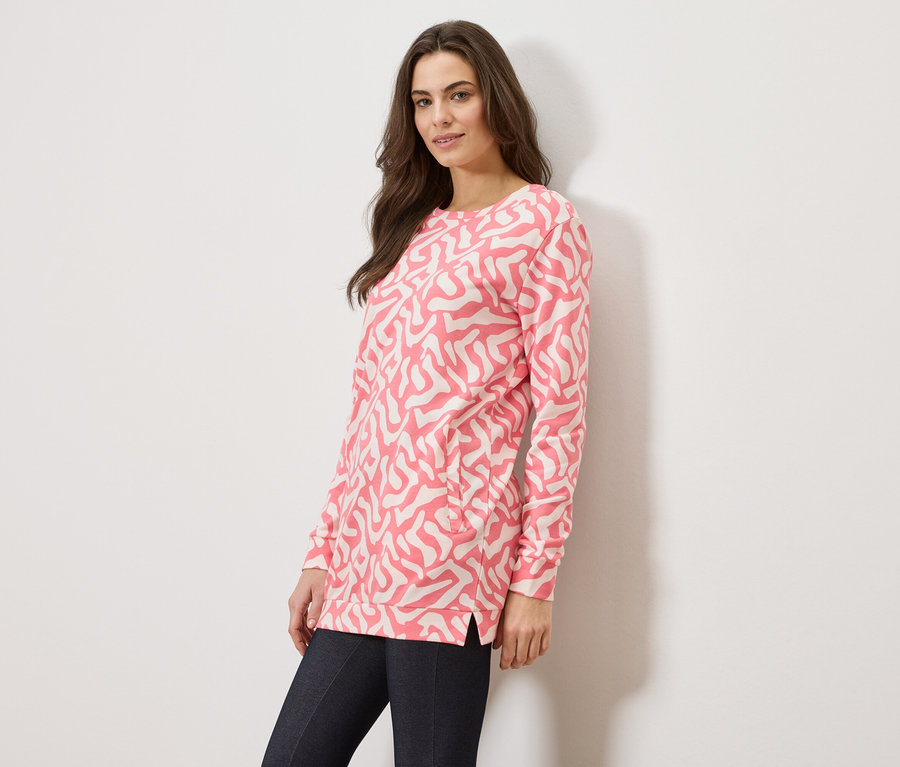 Nő visel rózsaszín, absztrakt mintás pulóvert és sötét farmert fehér háttér előtt.