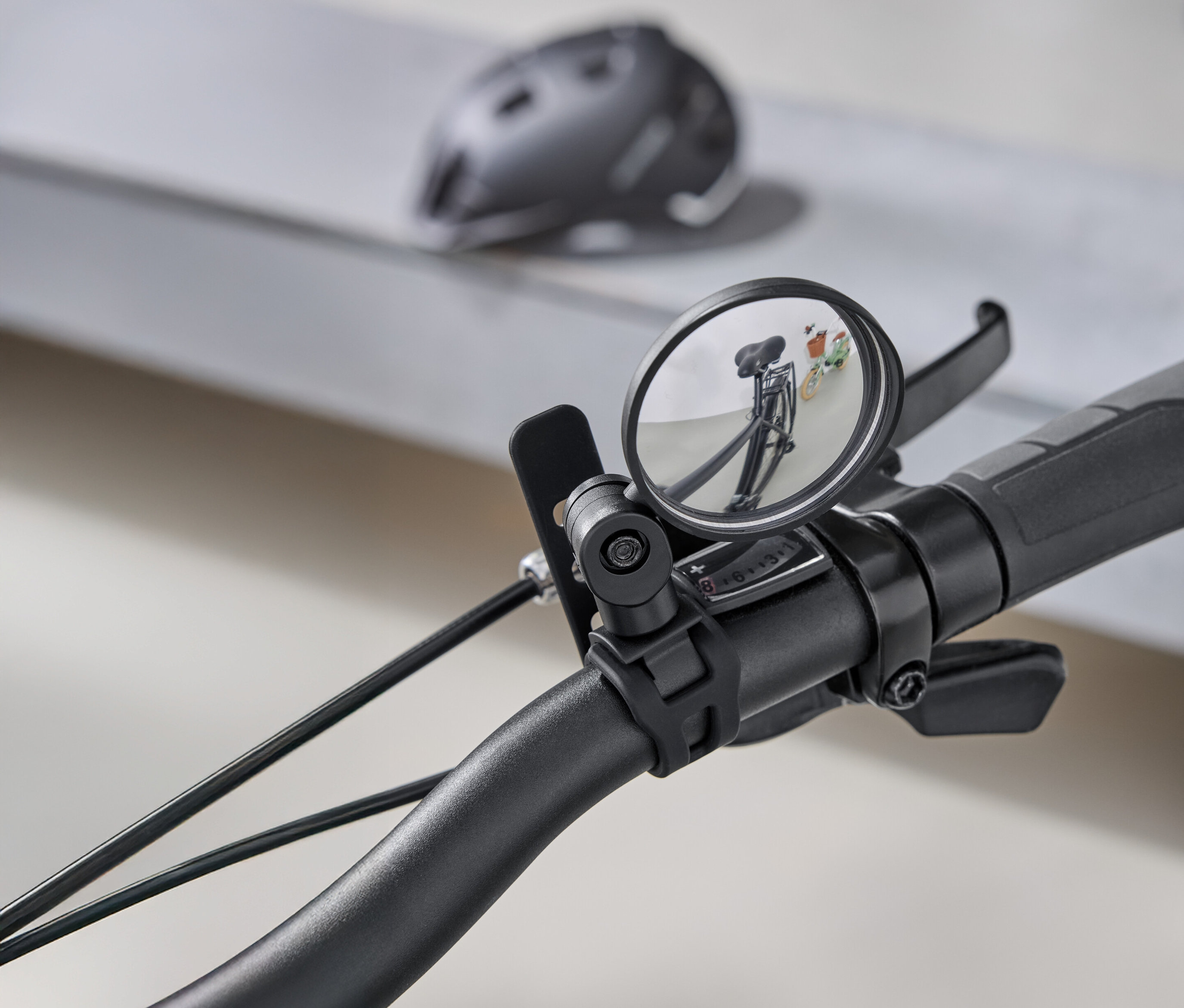 Egy kerékpár kormányának közeli képe, kerek visszapillantó tükörrel, amely a mögötte lévő utat tükrözi. A háttérben egy szürke kerékpáros sisak látható.