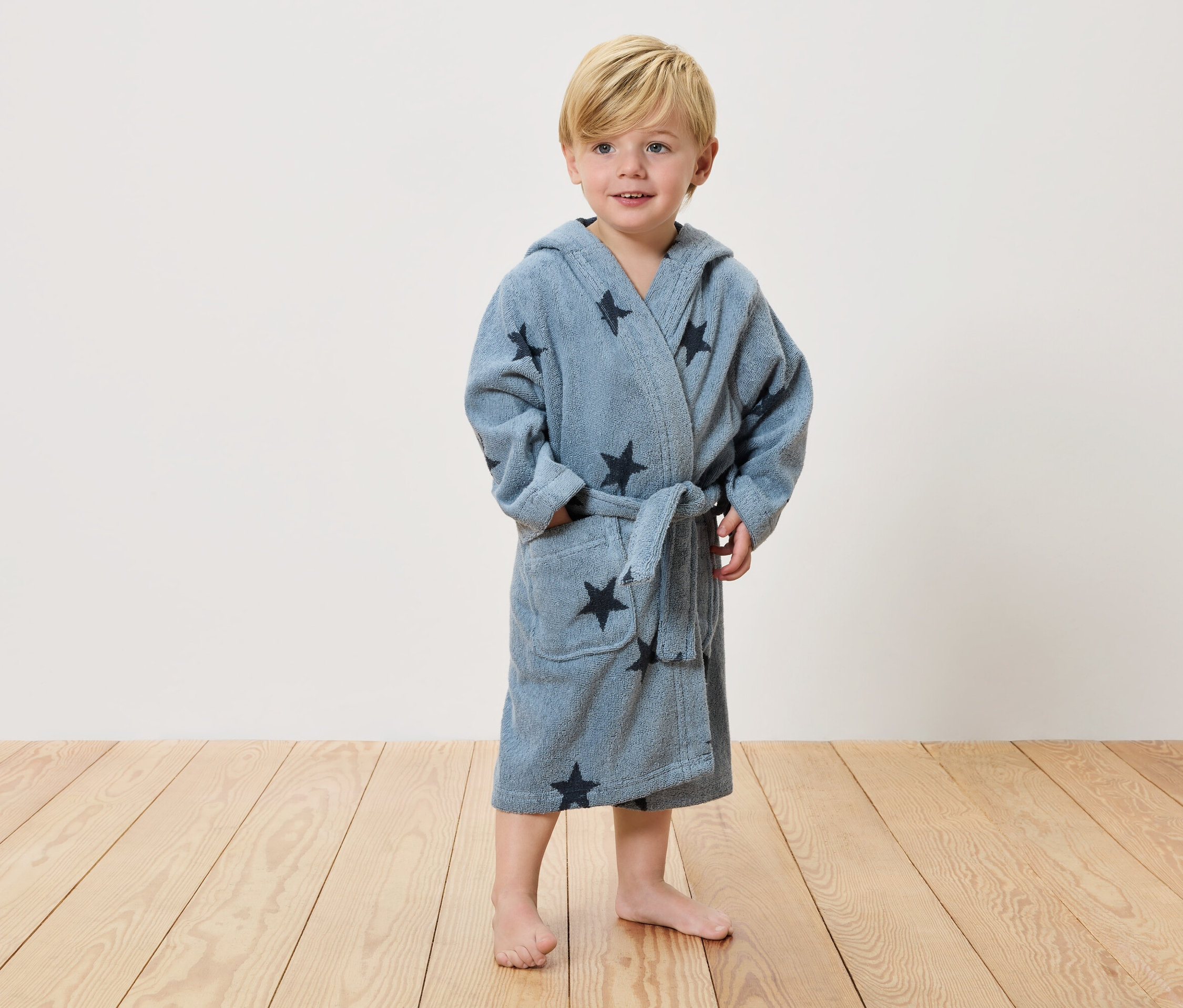 Egy szőke hajú kisfiú áll mezítláb egy fapadlón, világoskék, sötét csillagokkal díszített köntöst visel.