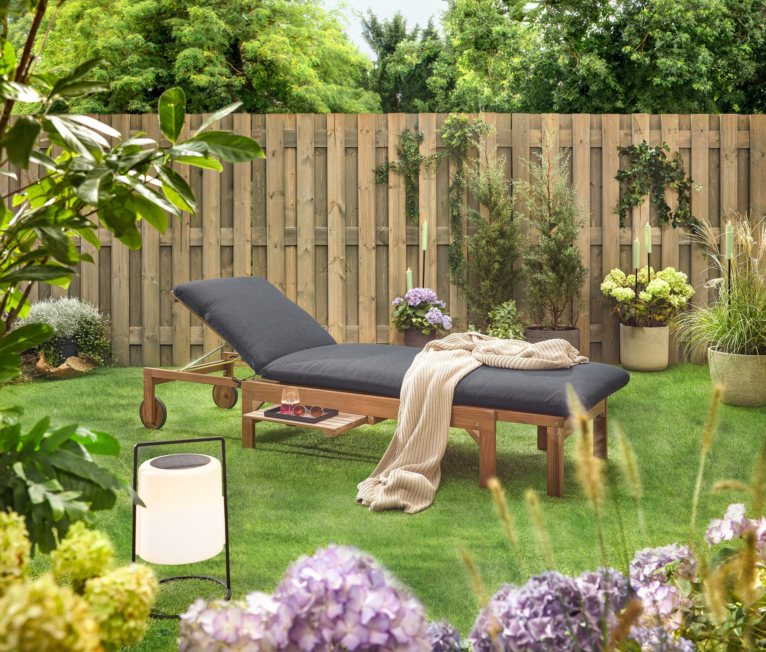 Kerti jelenet fa nyugággyal, fekete kárpitozással, takaróval, itallal, napszemüveggel és lámpással a fa kerítés előtti füvön.