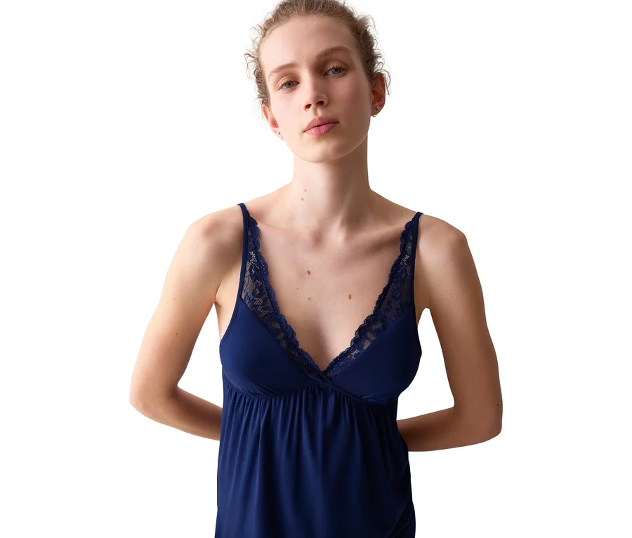 Egy nő sötétkék, csipkés szegélyű camisole-t visel, kezeit a háta mögött tartva áll és előre néz.