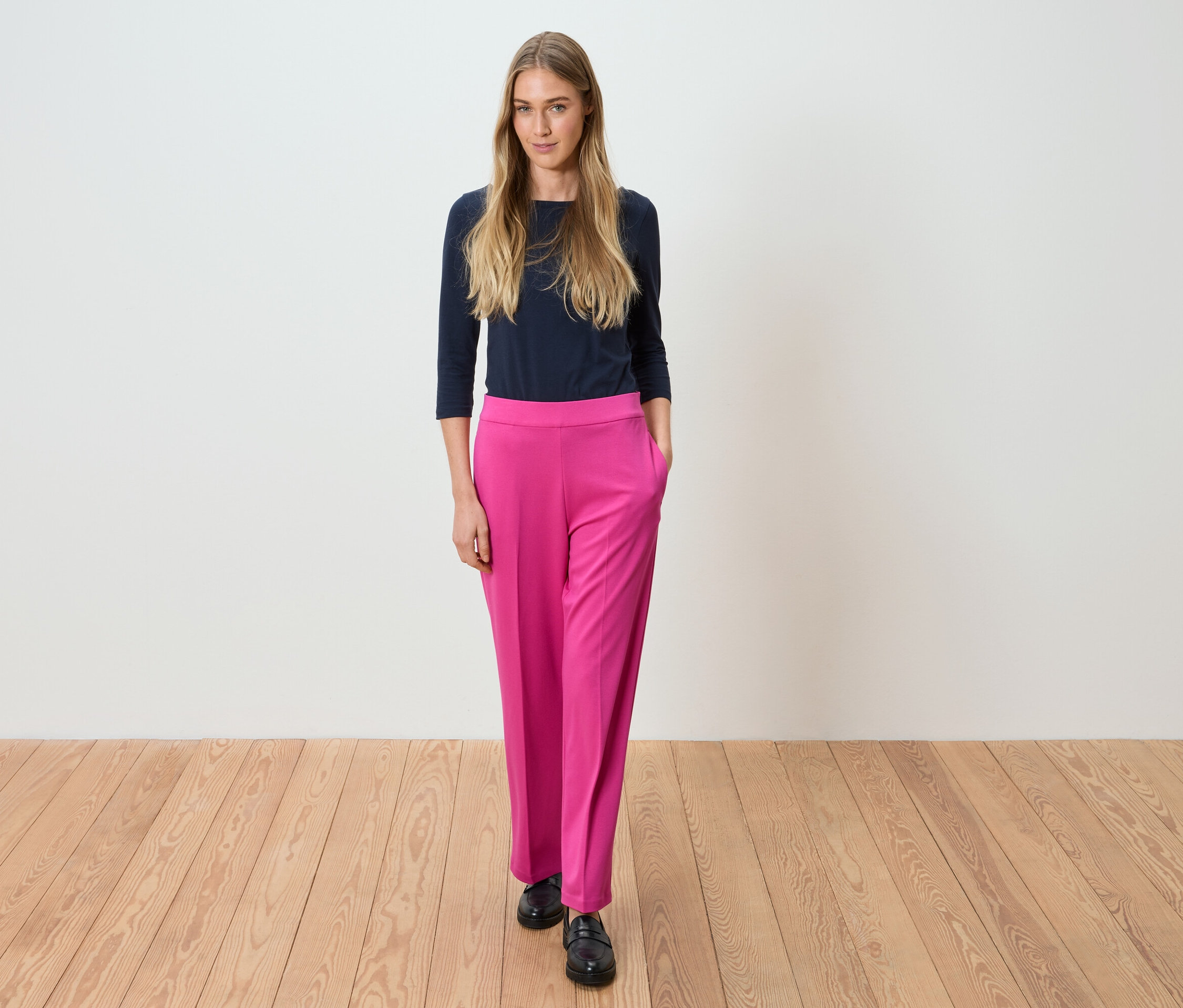 Nő áll sötétkék felsőben és rózsaszín nadrágban egy fehér fal előtt.