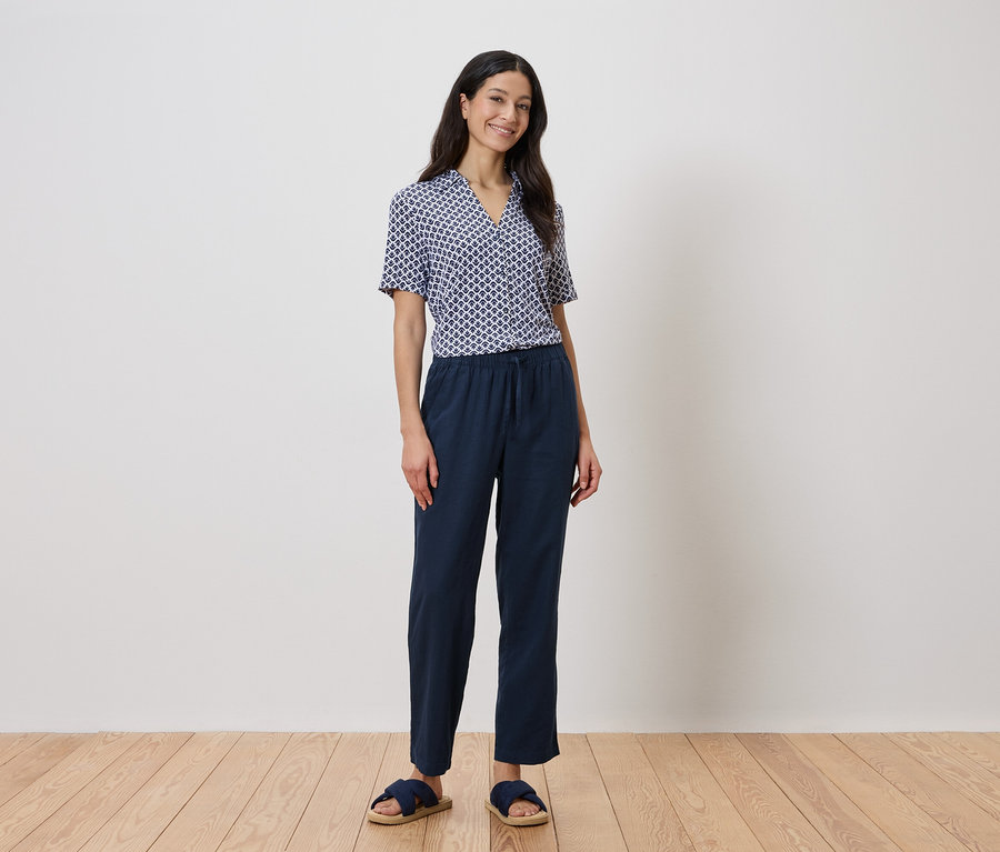 Egy nő áll a fal előtt, kék geometriai mintás inget és sötétkék nadrágot visel.