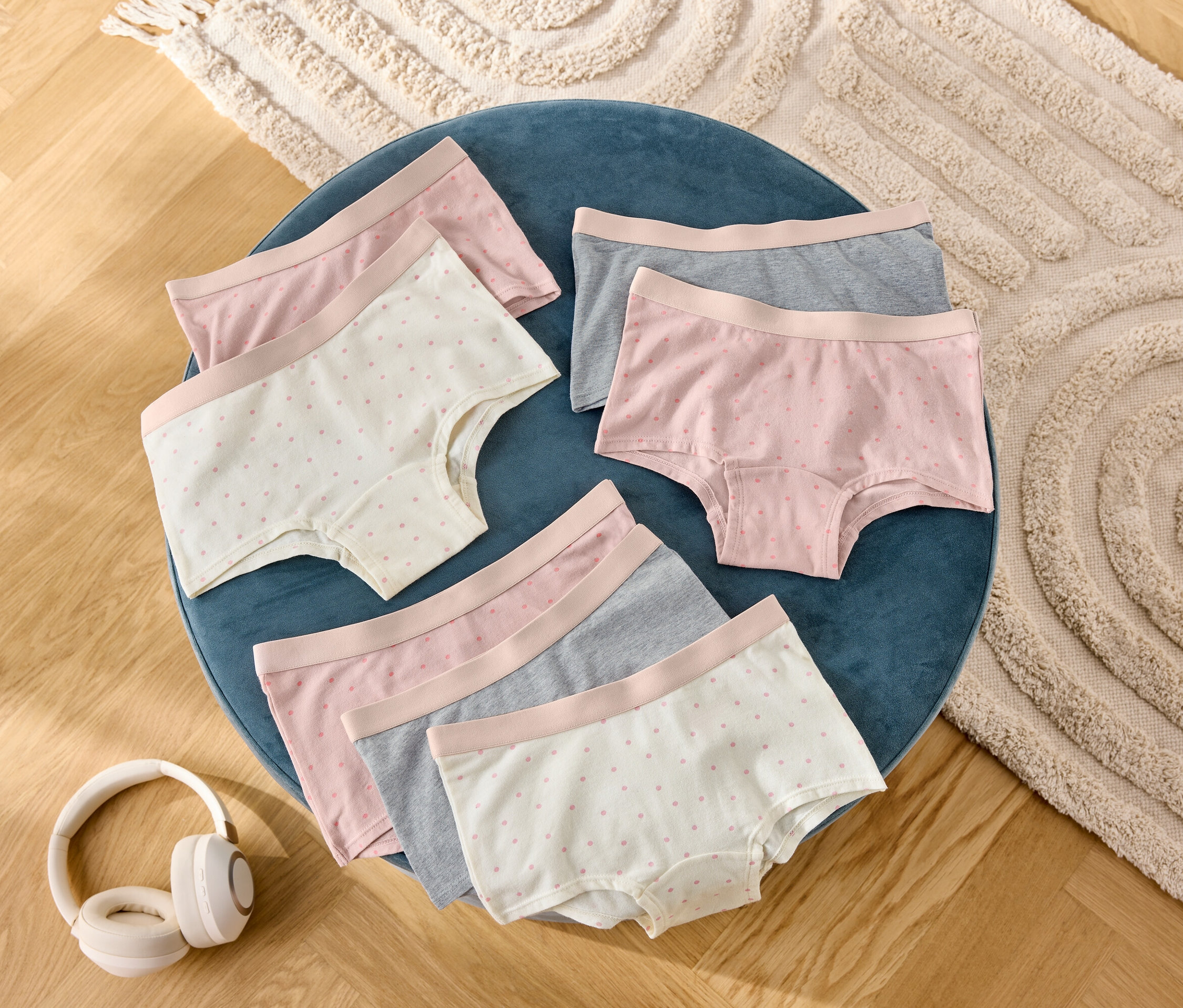 Egy kék zsámolyon hat női rövidnadrág fekszik szürke, rózsaszín és pöttyös színben, mellette fejhallgató.