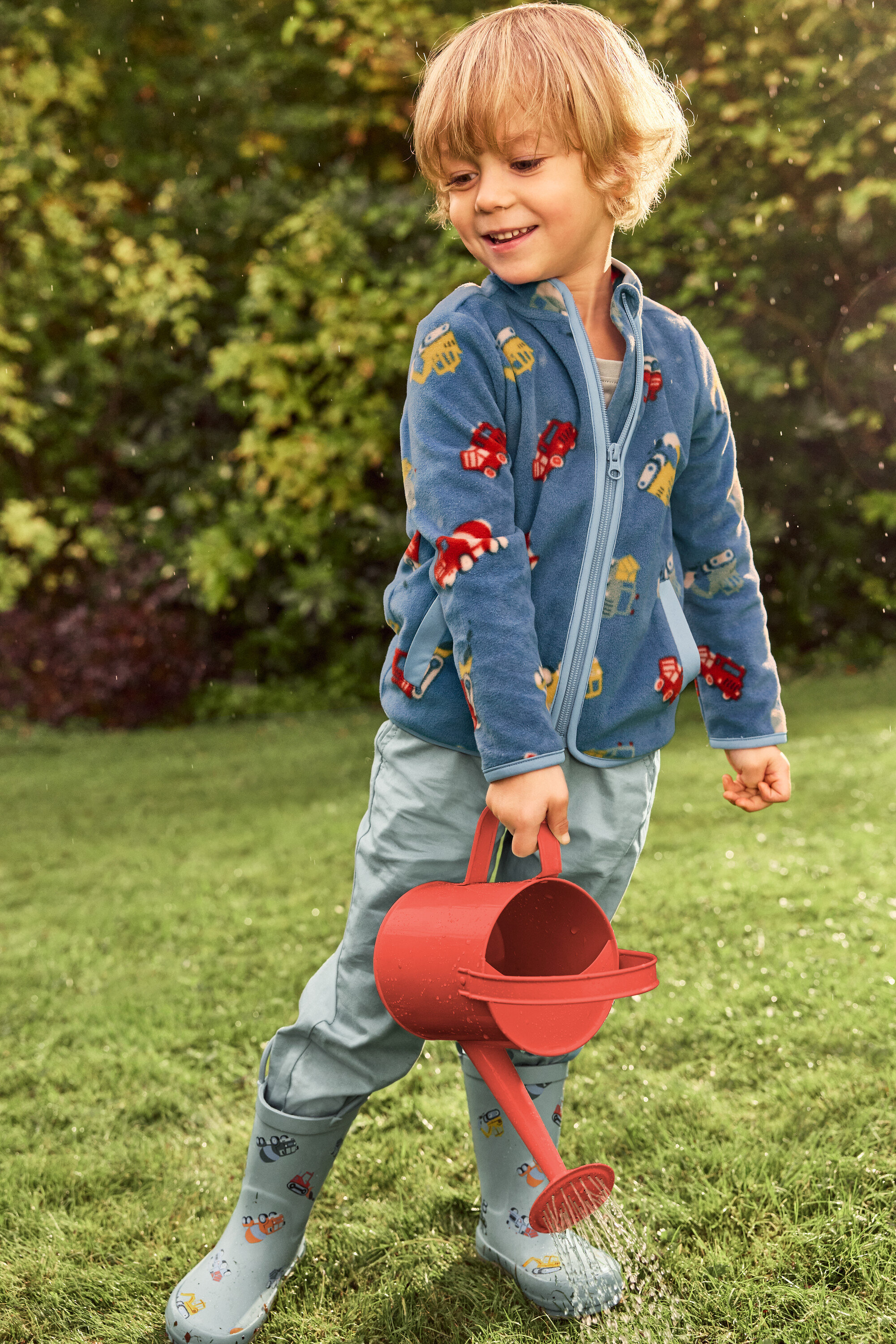 Egy mosolygó, szőke hajú fiú piros locsolókannával locsol egy zöld gyepet. Kék, járműmintás kabátot és szürke, szintén járműmintás gumicsizmát visel.