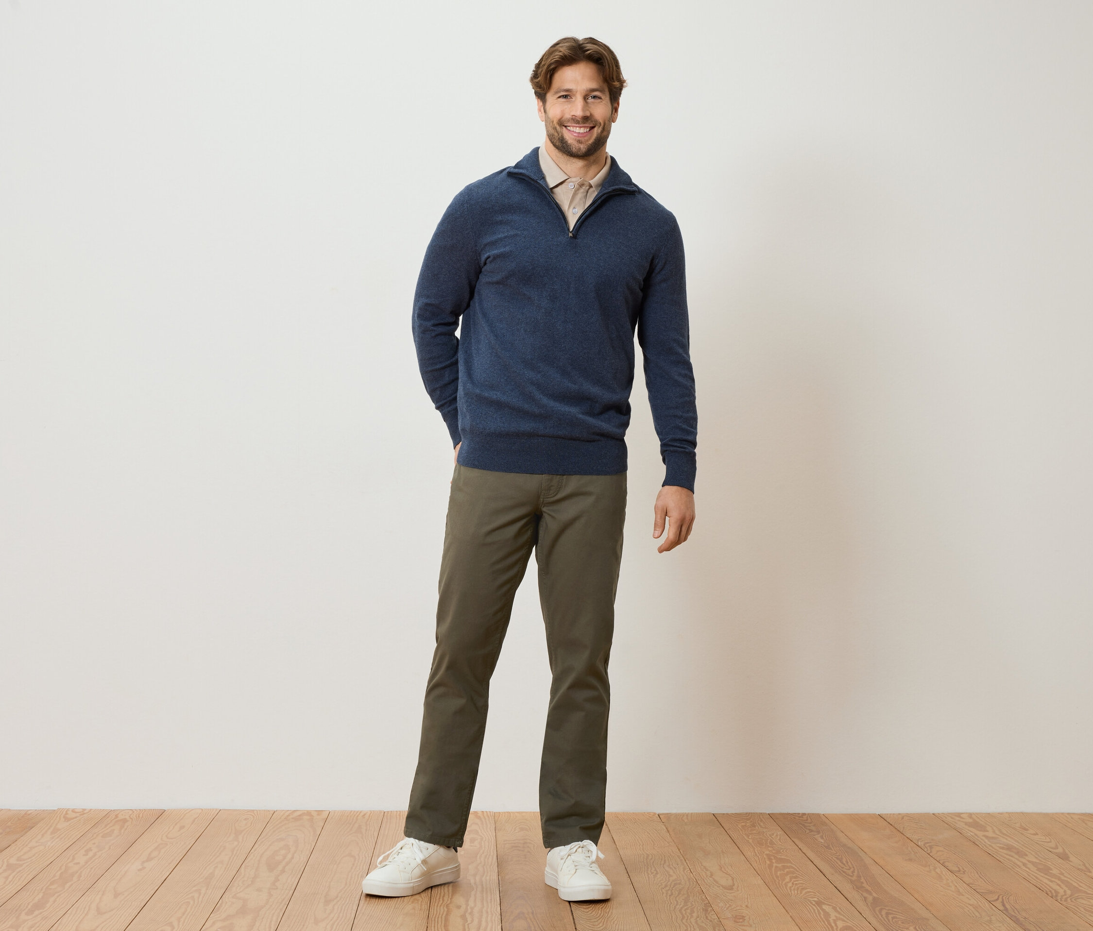 Egy férfi áll a fal előtt zsebre tett kézzel. Sötétkék pulóvert, világosbarna blúzt, khaki nadrágot és fehér tornacipőt visel.