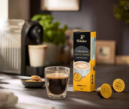 Az asztalon kávéfőző, egy pohár kávé és egy Caffè Crema mild – 120 db kávékapszula csomagolása látható.
