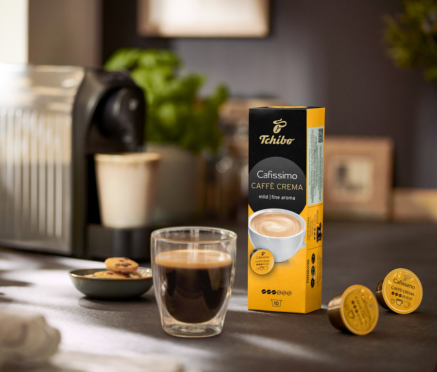 Az asztalon kávéfőző, egy pohár kávé és egy Caffè Crema mild – 120 db kávékapszula csomagolása látható.