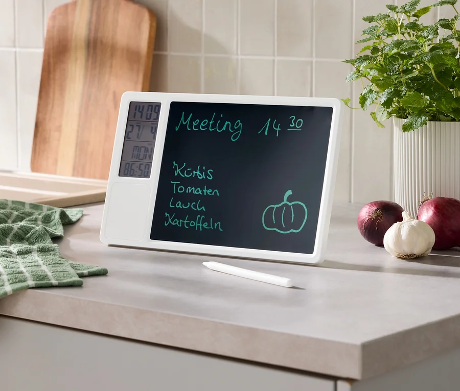 Fehér digitális emlékeztető tábla konyhapulton, rajta megbeszélés, bevásárlólista, idő, dátum és hőmérséklet. Mellette ceruza, zöld törölköző, fa vágódeszka, cserepes növény, vöröshagyma és fokhagyma.