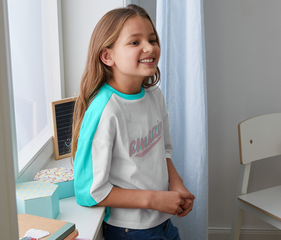 Egy mosolygó lány az ablakpárkányra támaszkodik, bio pamut pólót visel türkizkék ujjakkal.