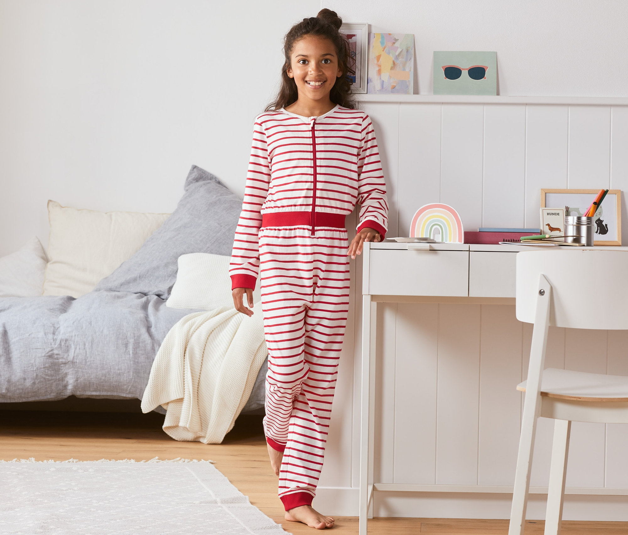 Egy lány egyrészes, csíkos, piros pizsamában áll a hálószobában.