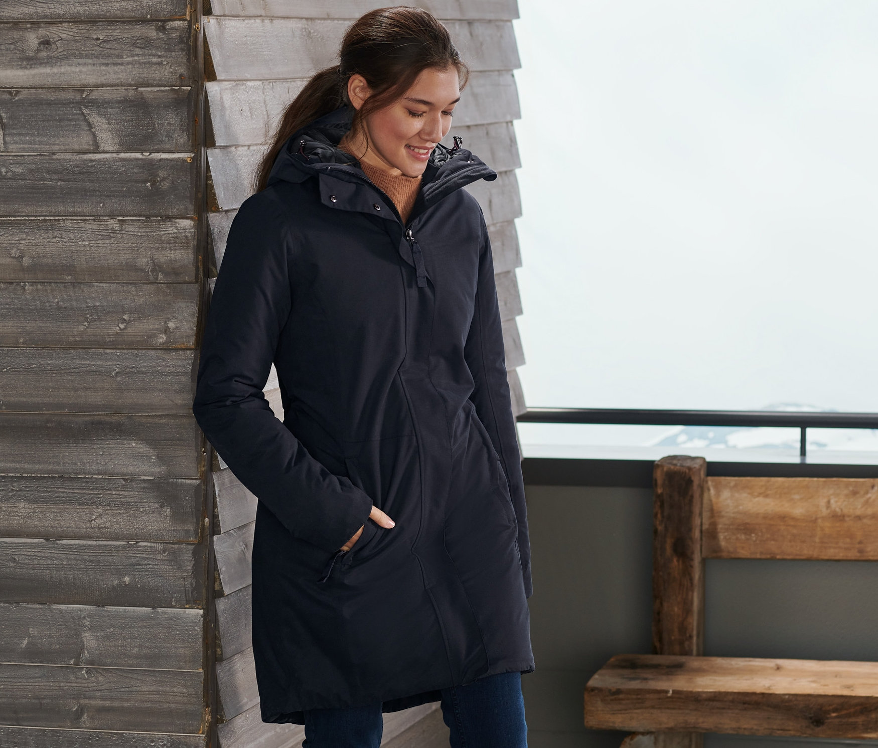 Egy nő a fal mellett áll sílécekkel, és sötétkék termokabátot visel.