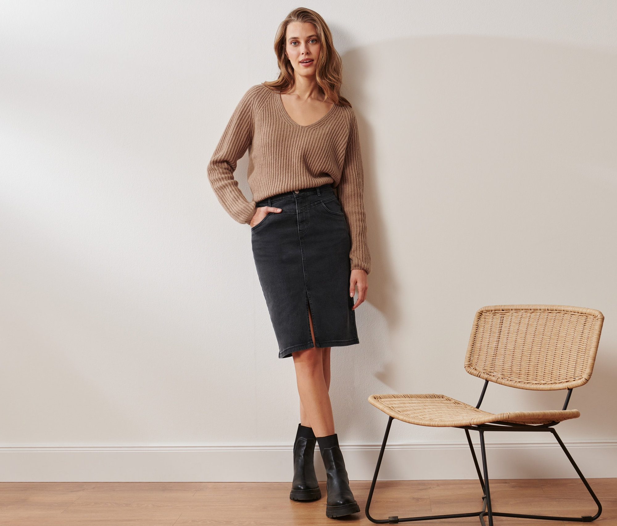 Egy nő pózol egy bézs színű, kötött pulóverben és egy fekete farmer ceruzaszoknyában. Mellette egy rattan szék áll.