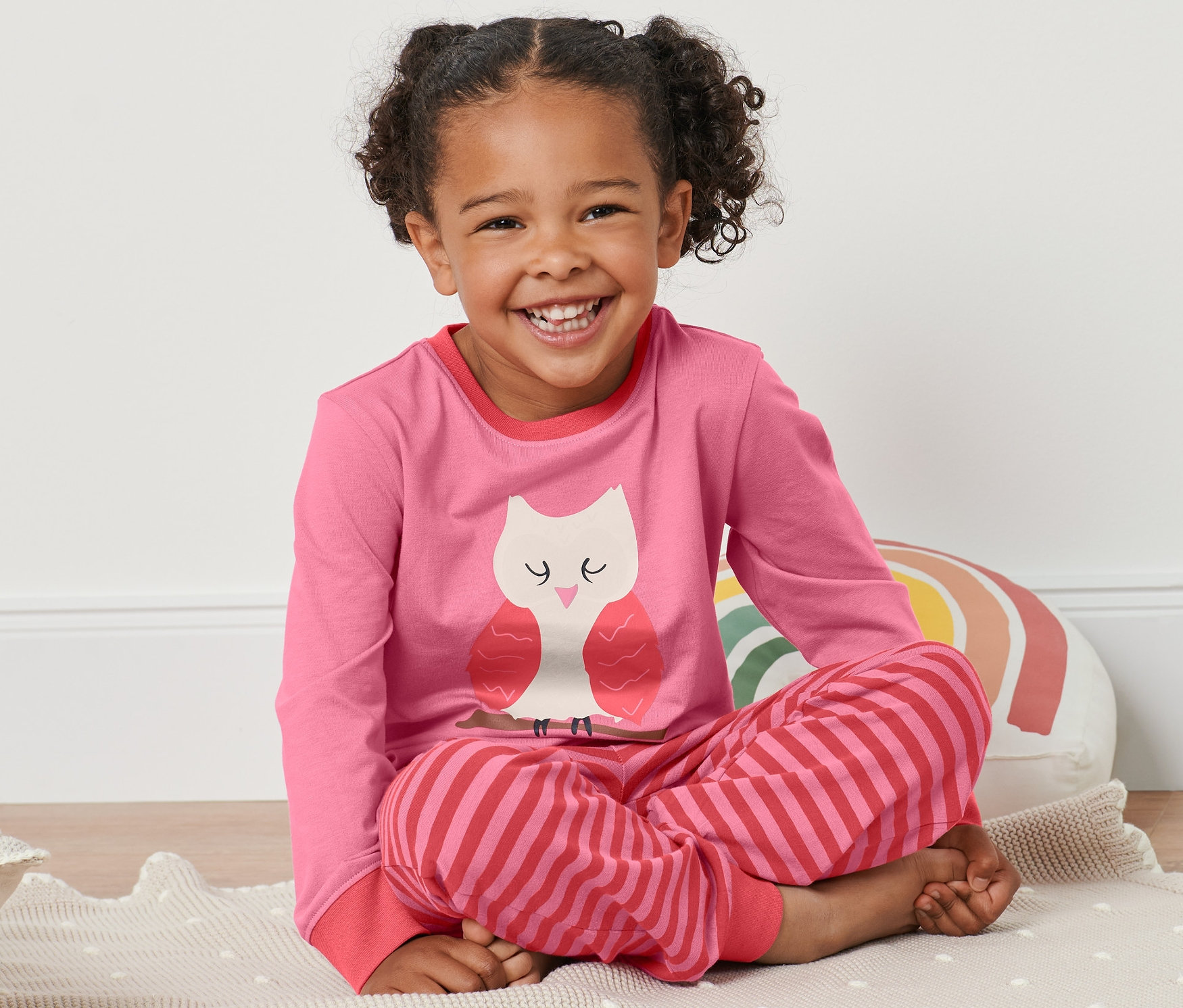 Kislány ül egy szőnyegen rózsaszín-piros, baglyos pizsamában.