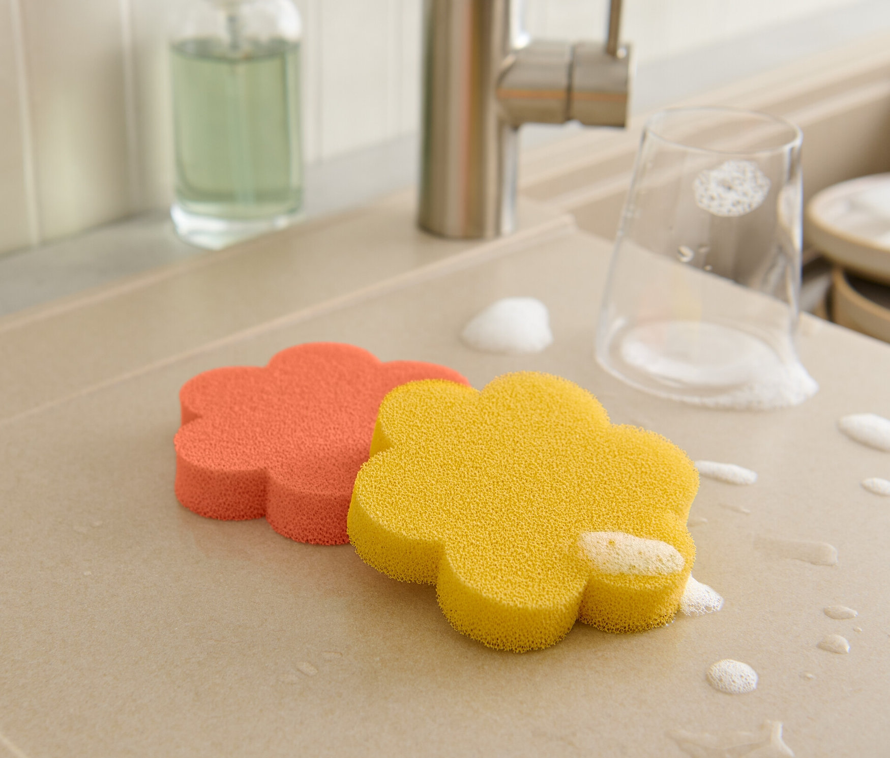 A csepegtetőtálcán narancssárga és sárga virág alakú szilikon szivacsok, egy pohár és edények vannak habbal borítva.