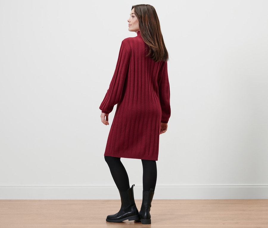 Nő pózol bordázott kötött ruhában, fekete leggingsben és fekete csizmában.