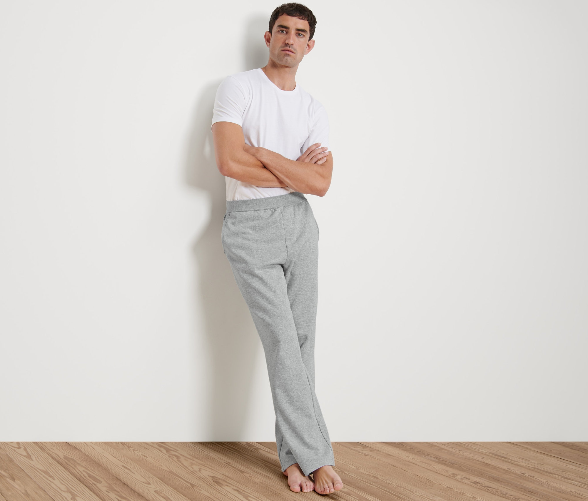 Férfi a falnak dőlve, fehér pólót és szürke férfi szabadidőnadrágot visel.
