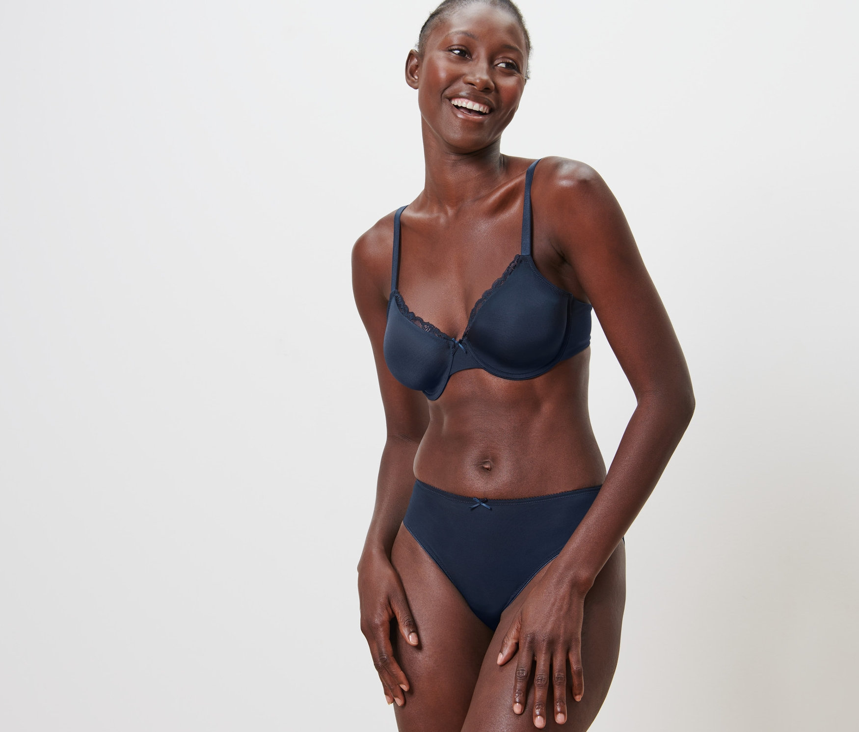 Mosolygó nő merevítős melltartót és alsóneműt visel, fehér háttér előtt pózol.
