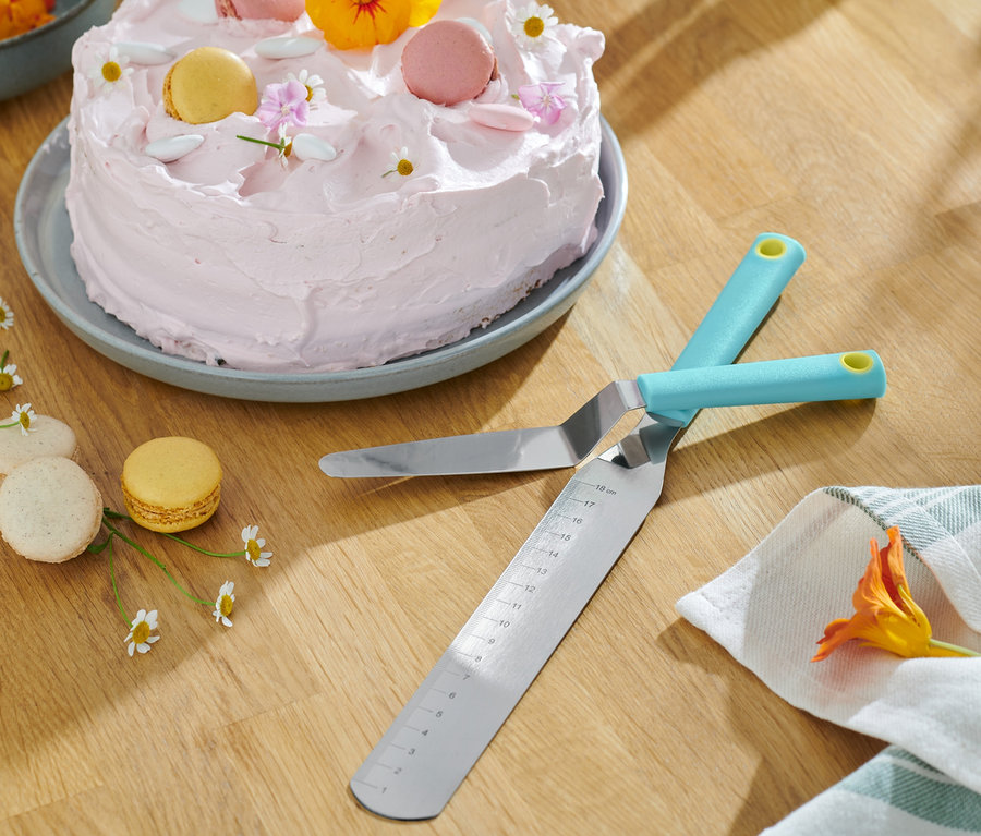 Két egymást keresztező 2 simító spatula egy faasztalon fekszik egy rózsaszín cukormázzal és makaronokkal díszített sütemény mellett.