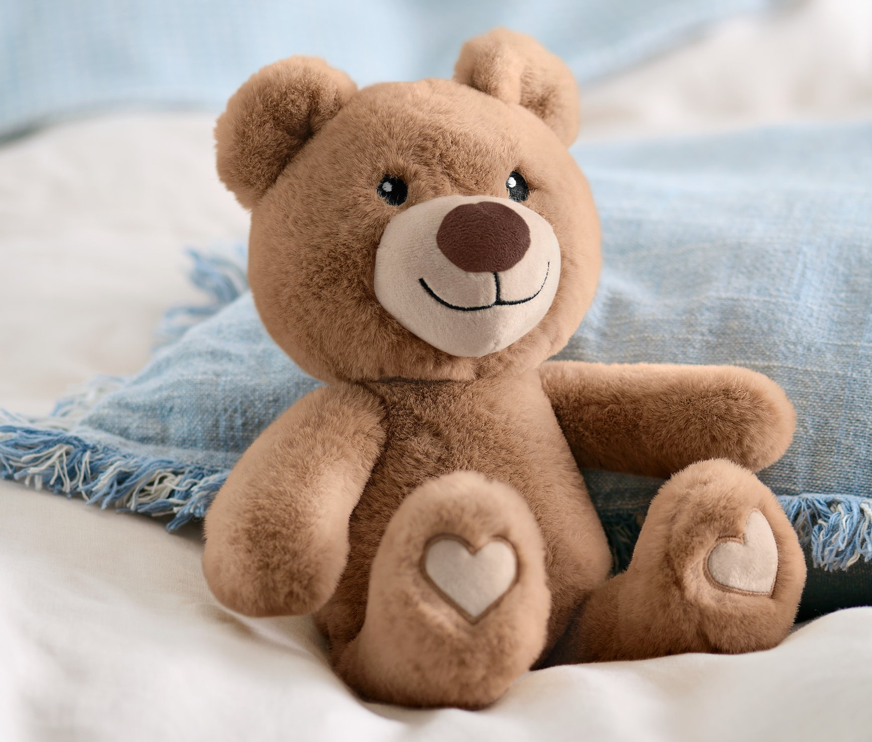 Egy barna Plüssmackó ül fehér ágyneműn és egy kék párnán.