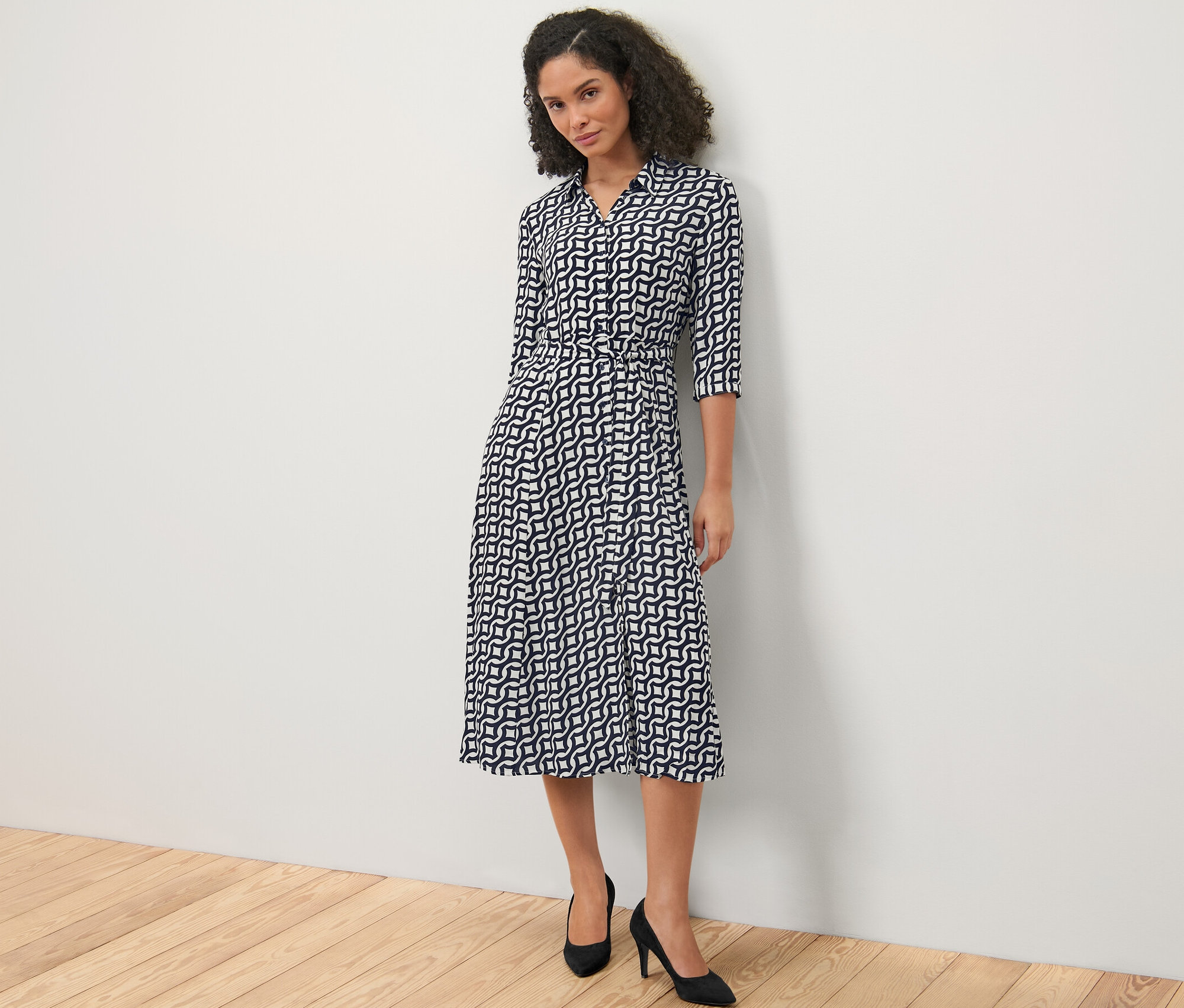 A fehér fal előtt álló nő sötétkék/krémfehér mintás blúzruhát és fekete magassarkút visel.