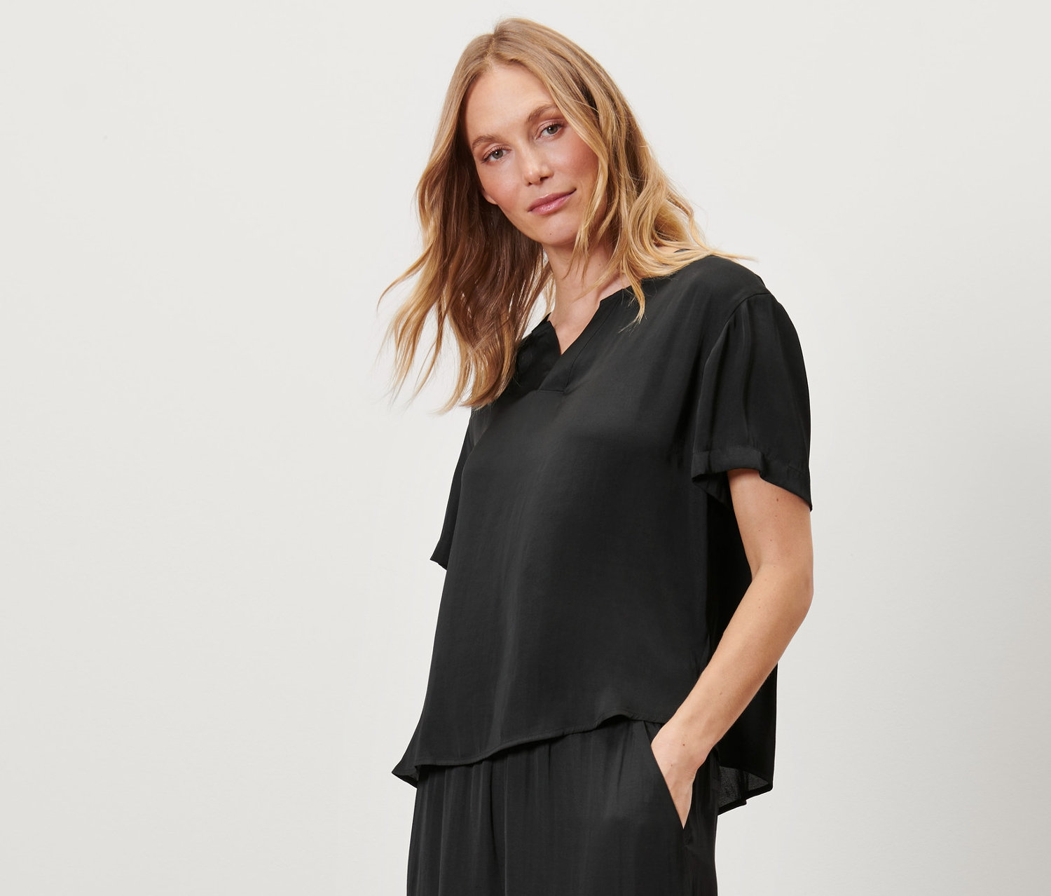 Egy nő áll, kezével a zsebében, fekete szatén tunikablúzt visel.