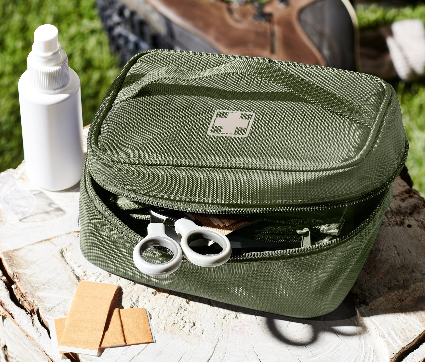 Egy zöld gyógyszeres táska fehér kereszttel nyitva fekszik egy fatönkön egy fehér pumpás palack és sebtapaszok mellett.