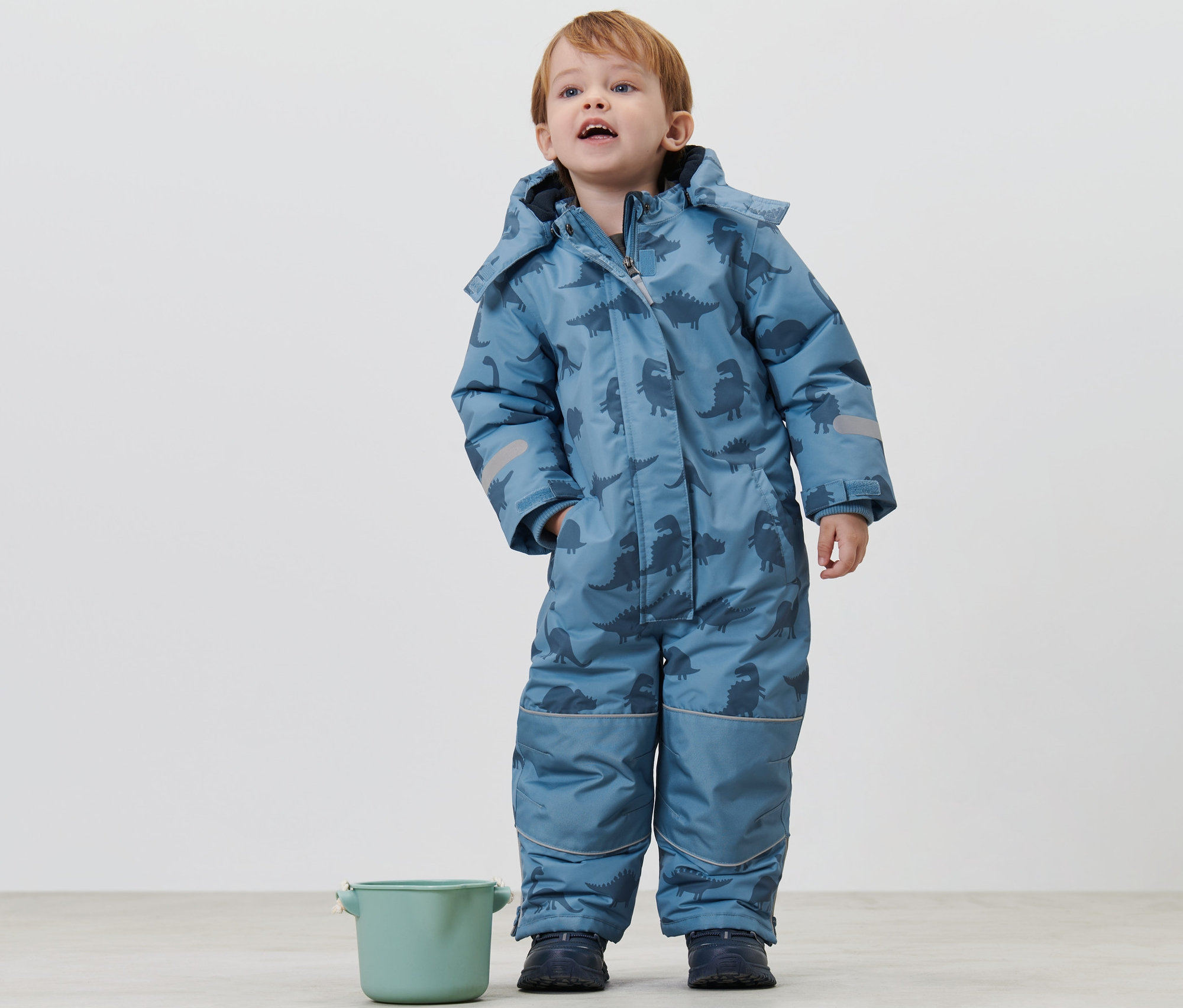 Fiúgyermek áll dínós gyerek téli kezeslábasban és felfelé néz. Mellette egy zöld vödör áll.