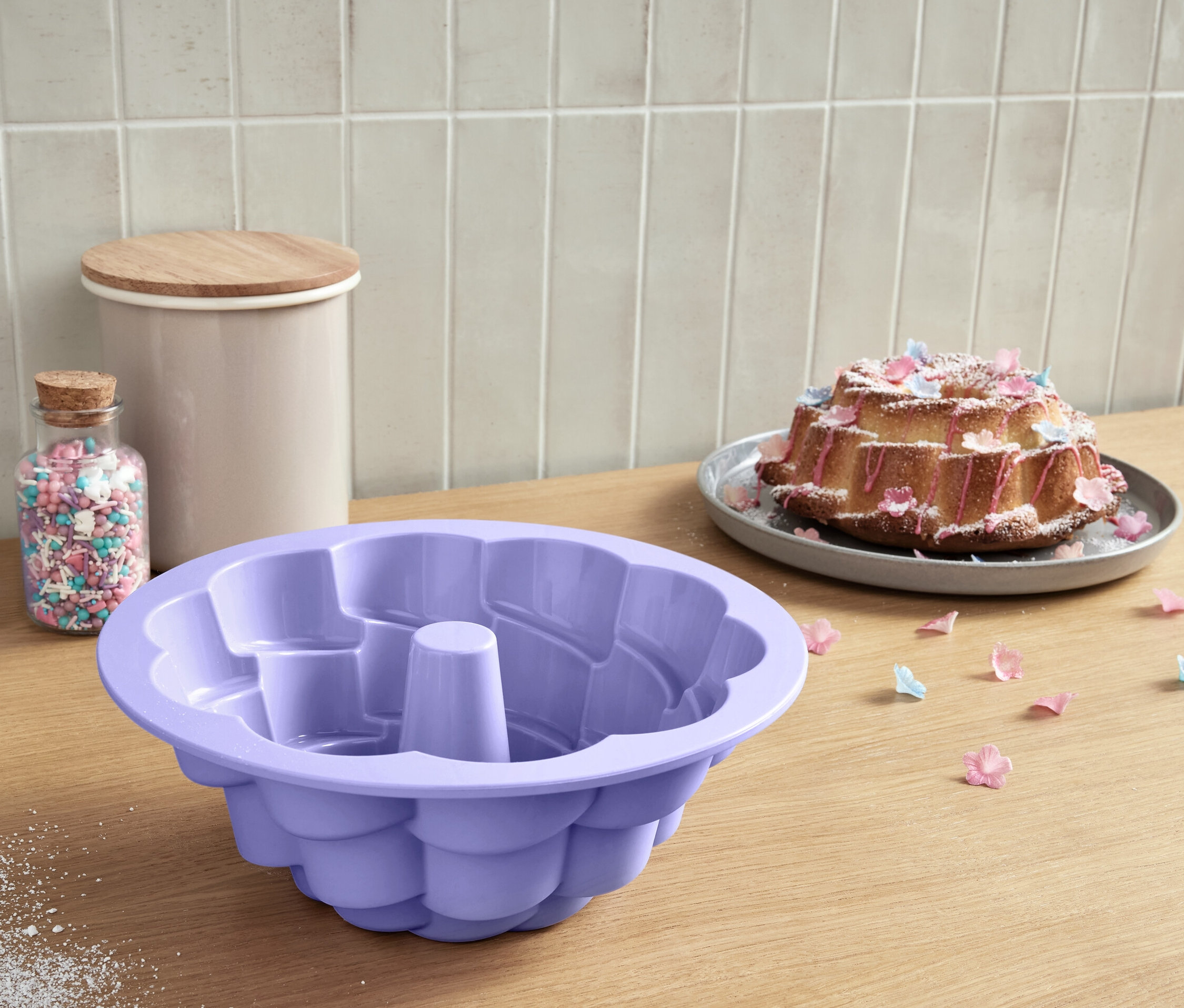 Lila, virág alakú szilikon kuglóf sütőforma egy fa asztalon, egy szórógyöngyökkel teli üveg mellett.