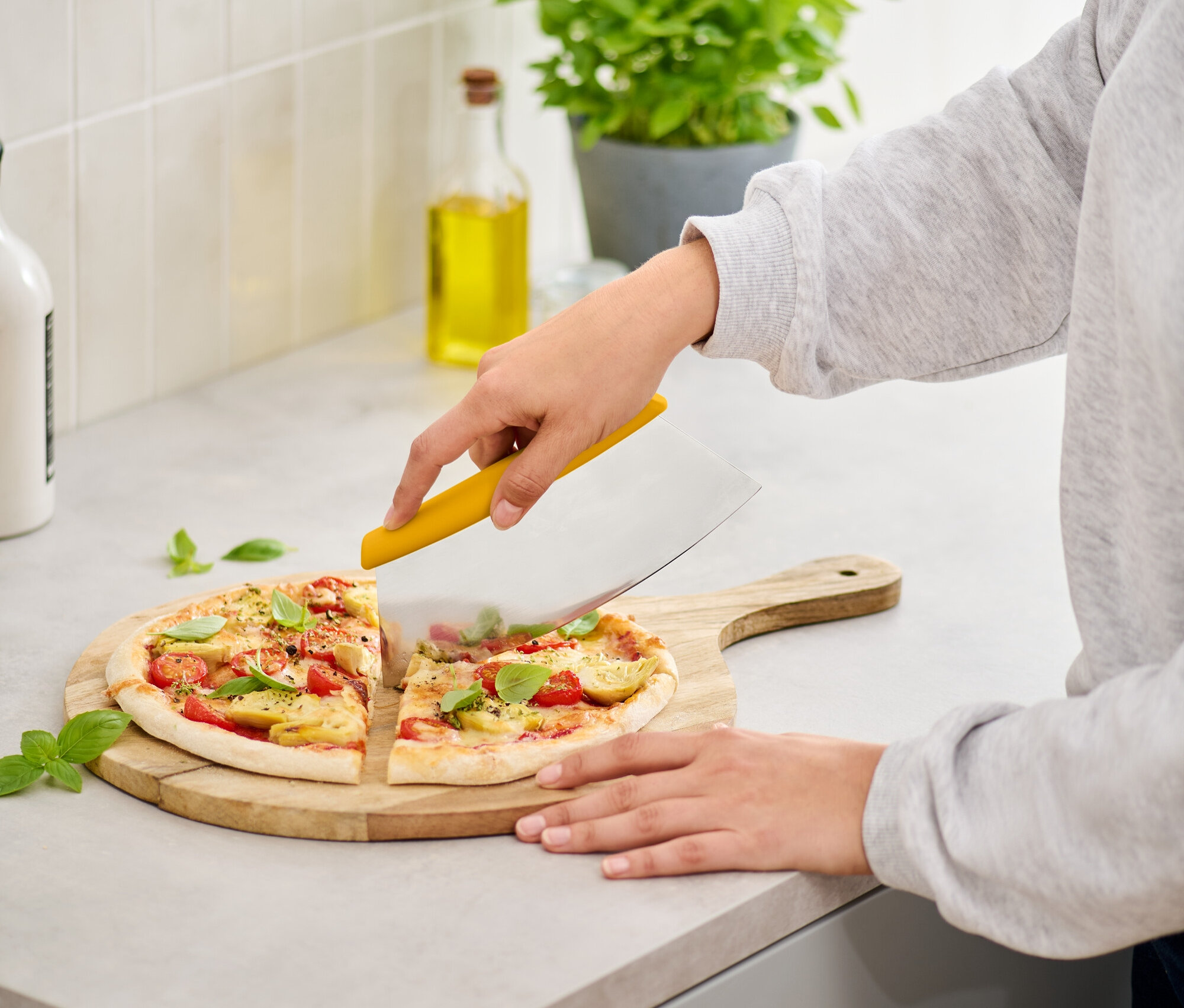 Nő paradicsomos, bazsalikomos pizzát vág fa vágódeszkán egy sárga pizzaszeletelővel.