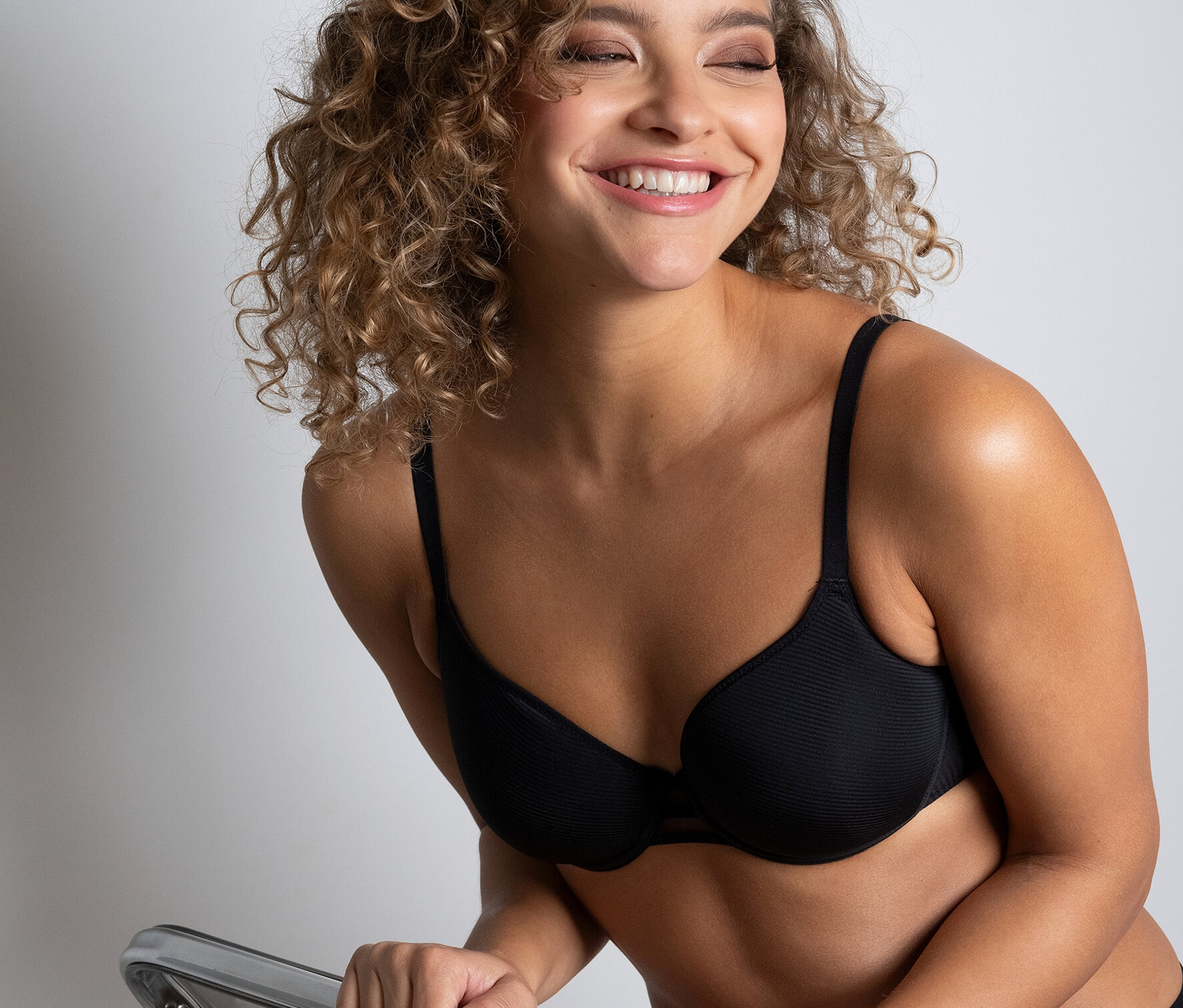 Mosolygó nő ül egy átlátszó széken, fekete Passionata »Nais« spacer melltartót visel.