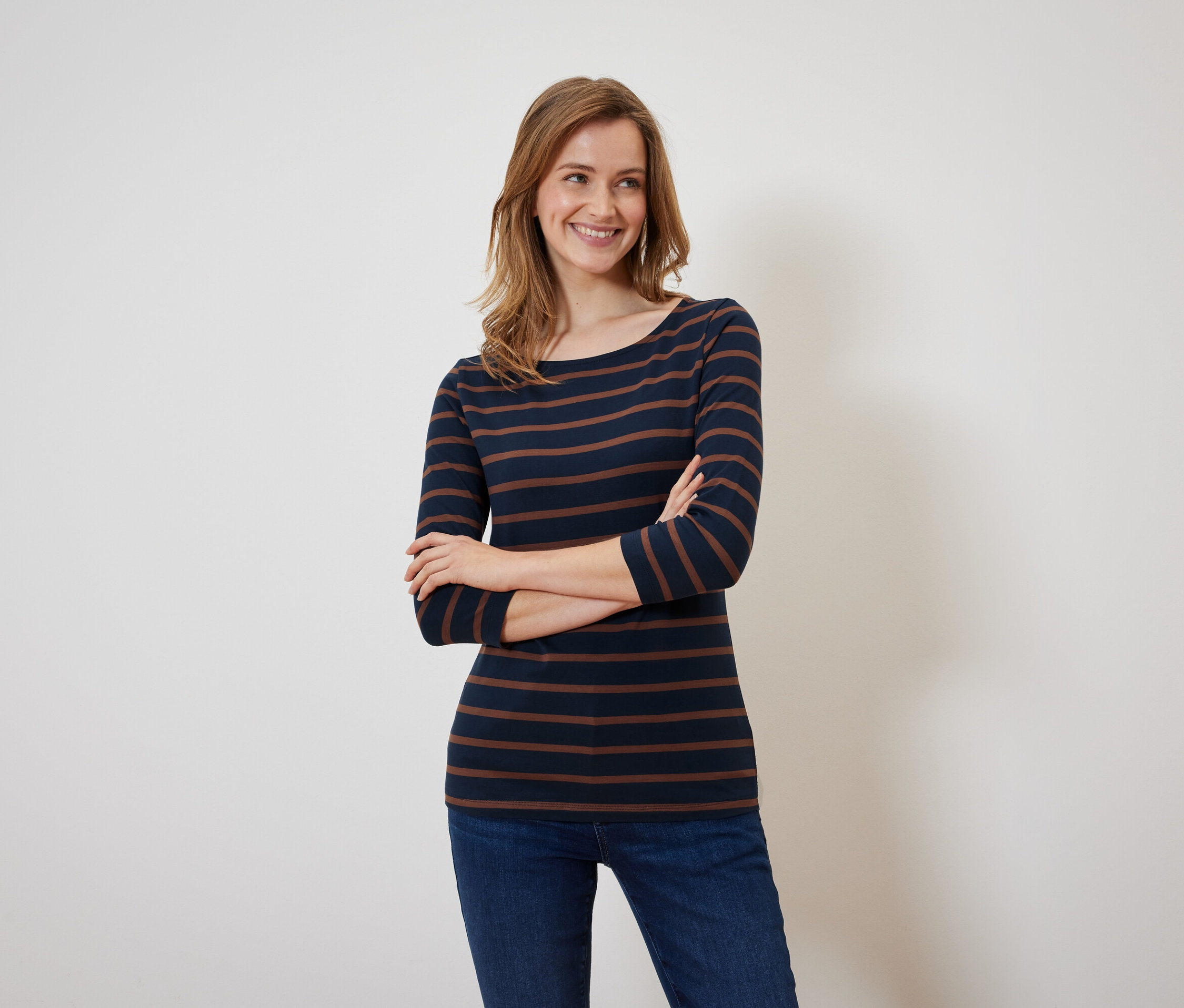 Egy mosolygó nő pózol karba tett kézzel, sötétkék és barna csíkos pólóban és farmerban.